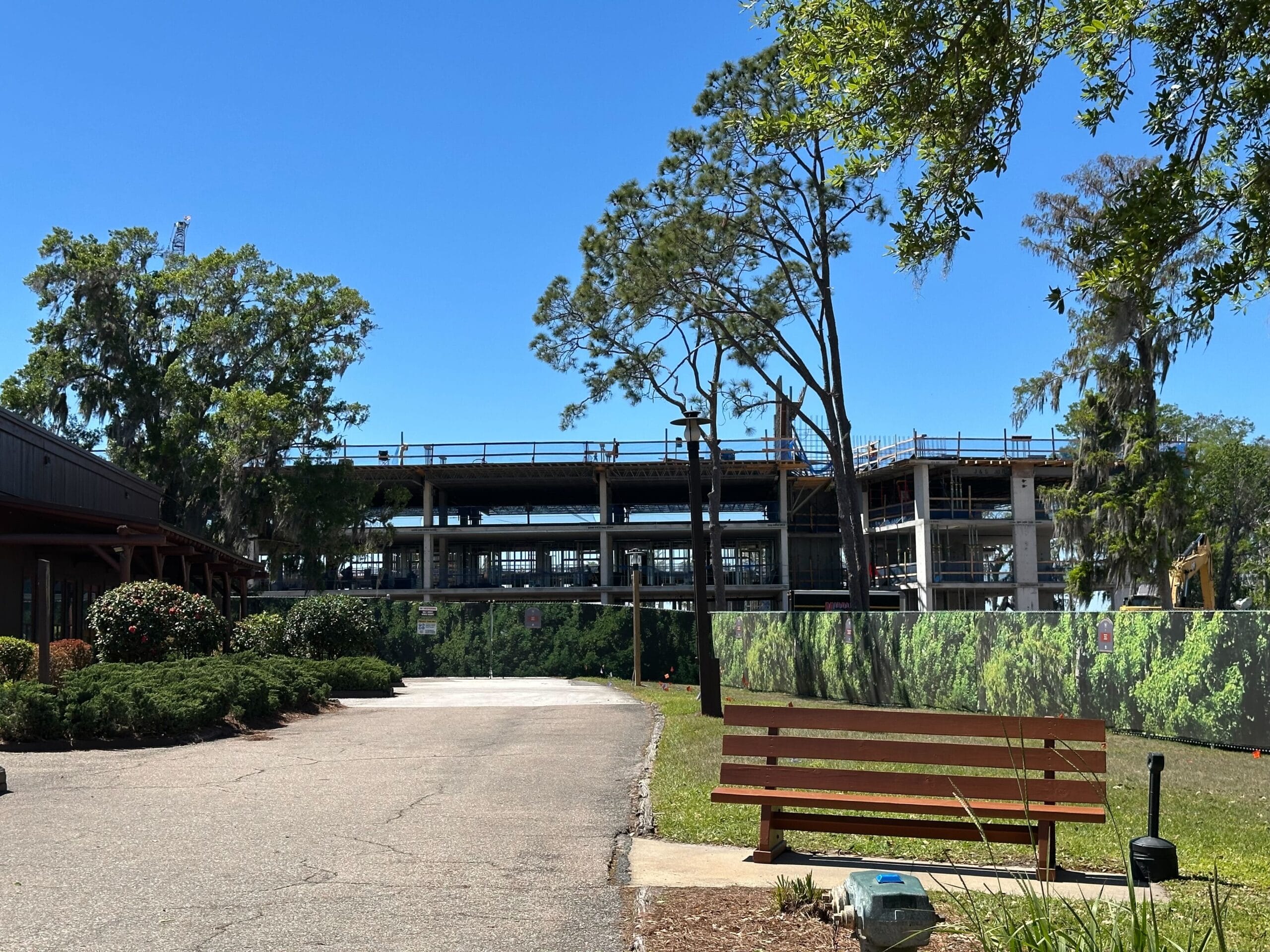 Construction site with a partially built structure surrounded by trees and a fence. A wooden bench is in the foreground, and the sky is clear and blue.