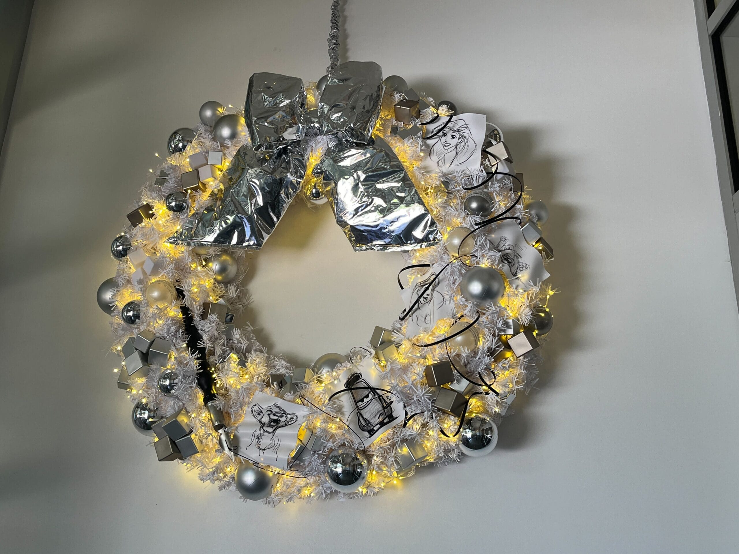 Silver and white wreath adorned with fairy lights, metallic ornaments, and a large bow hanging on a white wall.