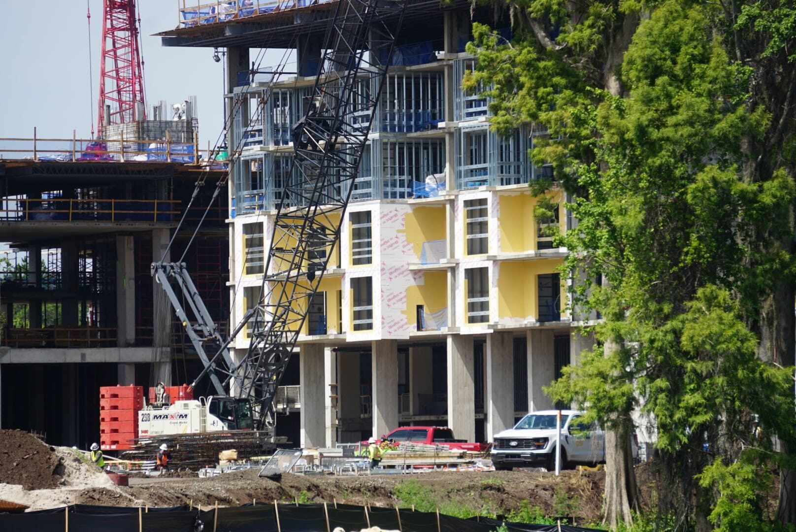 A construction site with cranes, workers, and a partially built multi-story building near trees.