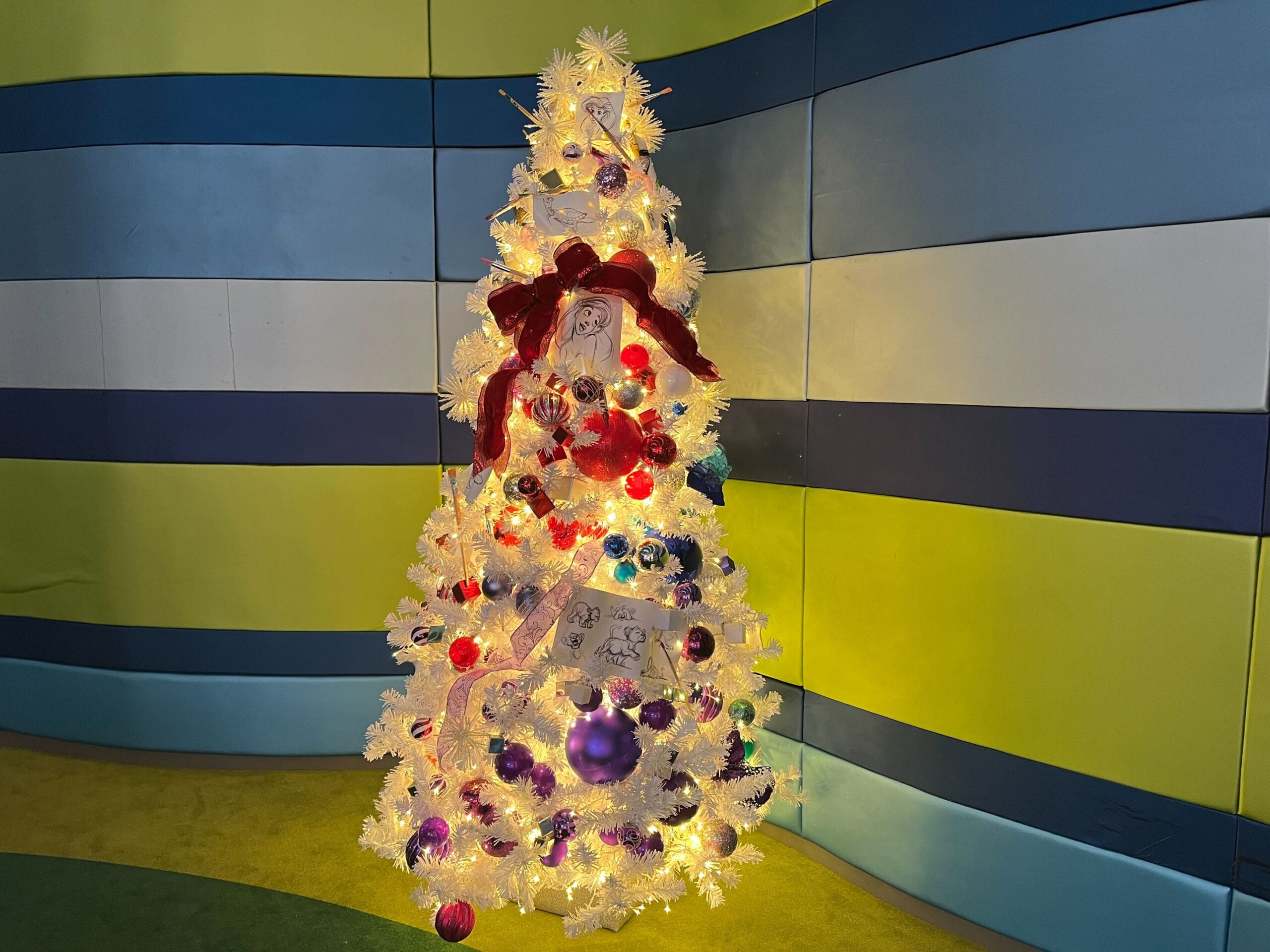A white Christmas tree with a red bow, multicolored ornaments, and lights stands against a striped blue and green wall.