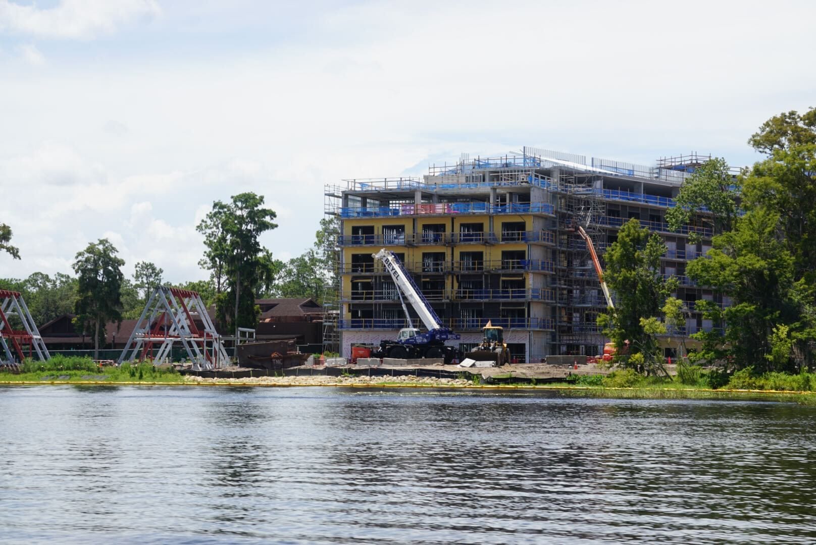 A multi-story building under construction by a lakeshore, with cranes and scaffolding visible.