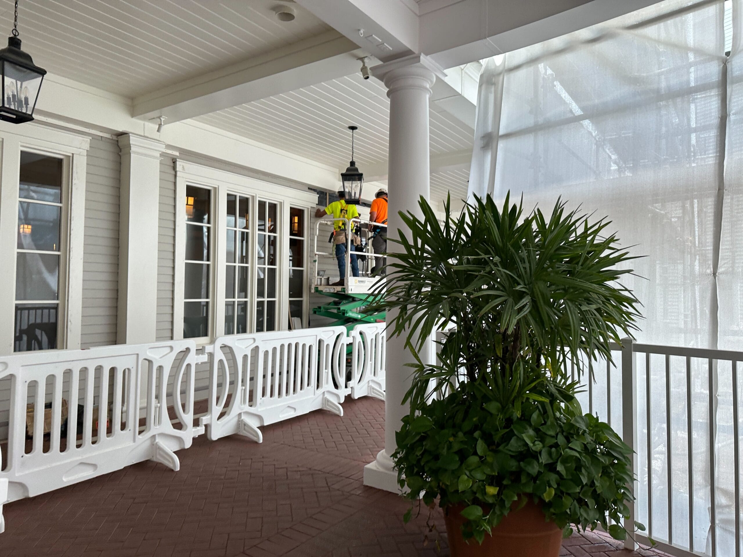 Workers on a lift perform maintenance near white barriers and potted plants on a covered porch area.