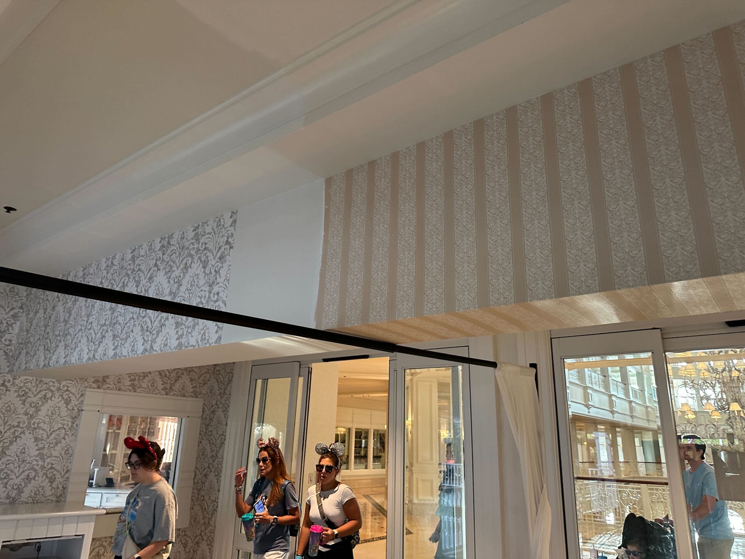 Four women wearing Mickey Mouse ears walk inside a room with patterned wallpaper and glass doors on a sunny day.