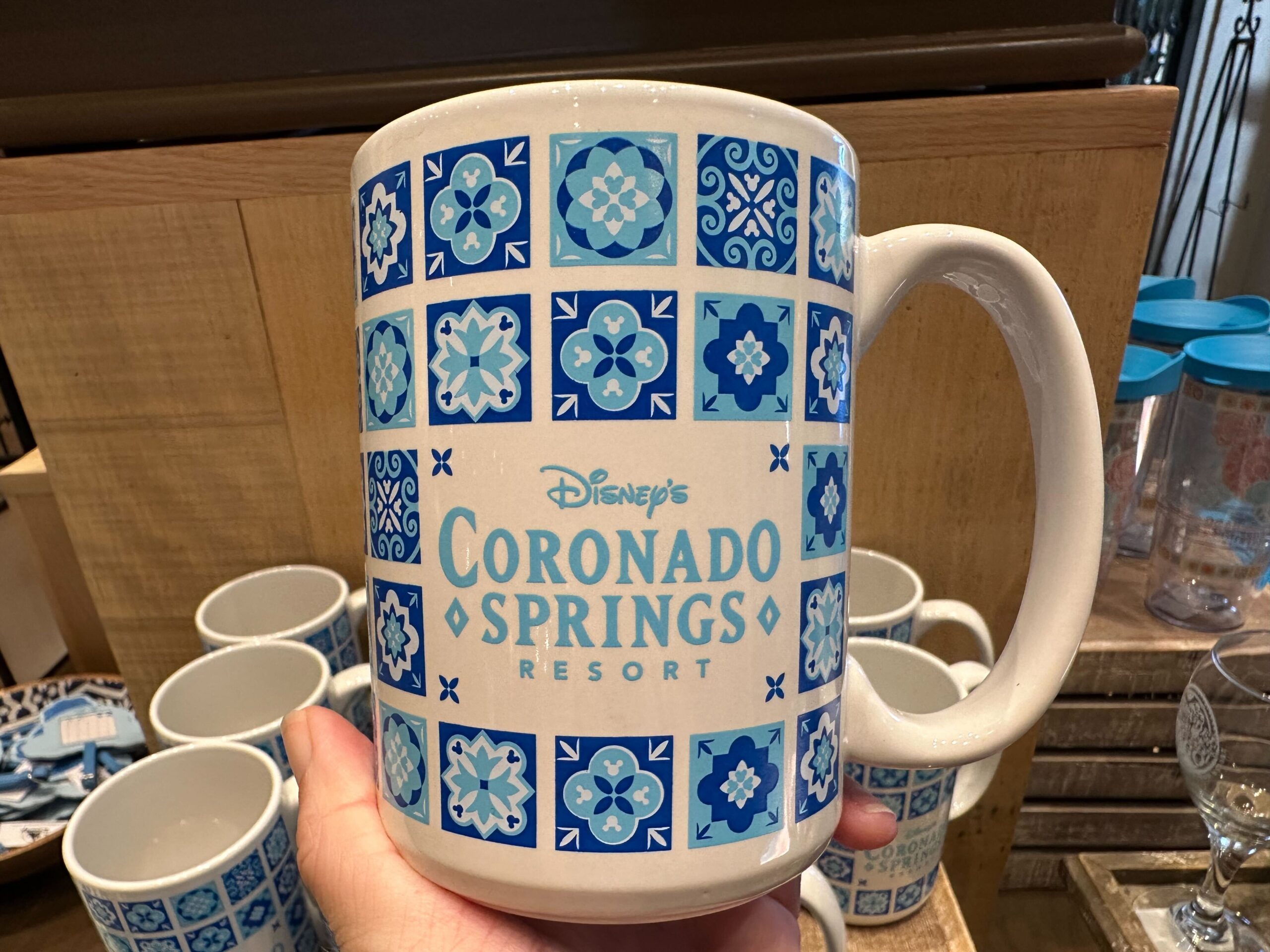 A hand holds a Coronado Springs Resort mug, showcasing its elegant blue tile pattern design. In the background, several similar mugs from the Coronado Springs merchandise line create an inviting display, perfect for adding charm to your collection or lug bag.