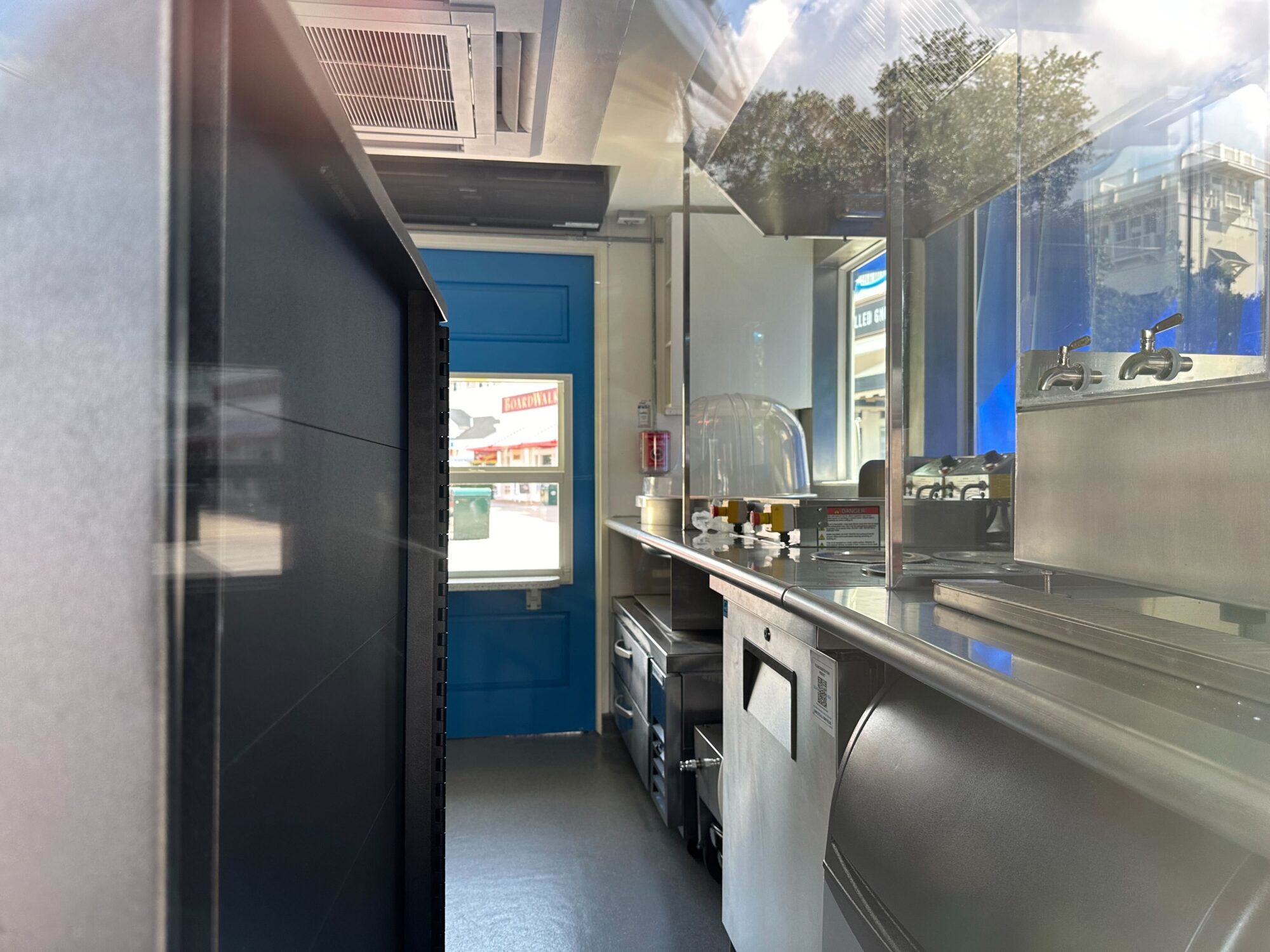 View inside a clean, modern food truck kitchen with stainless steel equipment, a blue door at the back, and various kitchen utensils arranged on the counter.