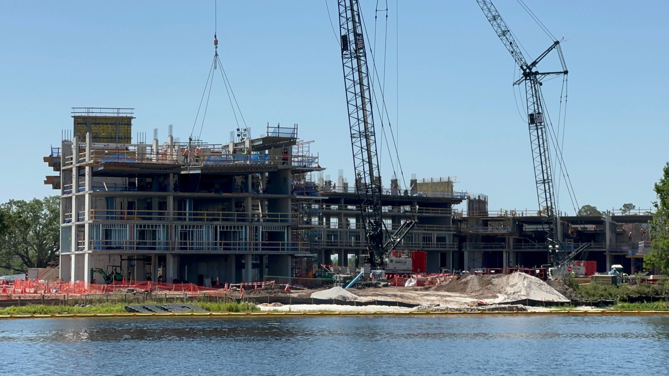 The construction site beside a serene lakeshore, reminiscent of a Disney fairytale, features cranes, scaffolding, and partially built structures basking under clear skies. This enchanting scene offers a magical construction update to behold.