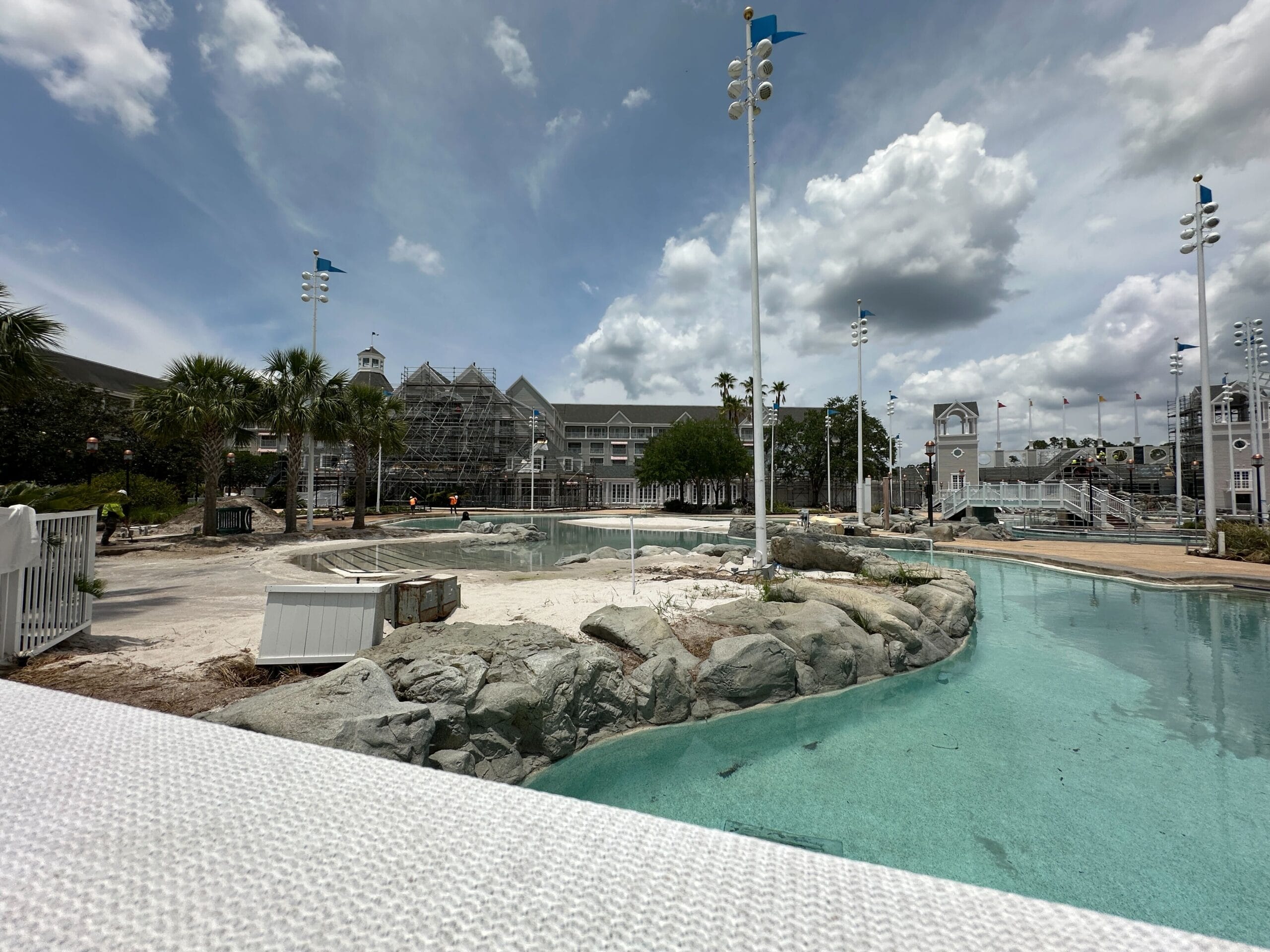 A resort pool area under construction with scaffolding, palm trees, rocks, and cloudy sky in the background.