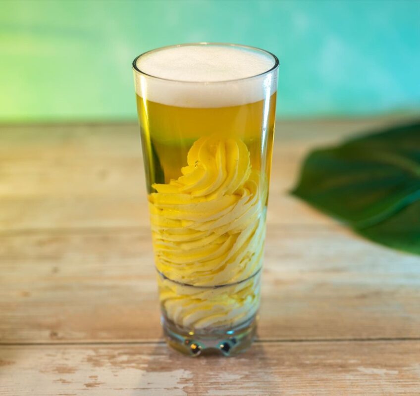 A tall glass filled with beer and a thick layer of foam on top, containing a swirl of butter at the bottom. The background features a blurred green and wooden surface with a leaf edge, capturing the essence of Typhoon Lagoon.