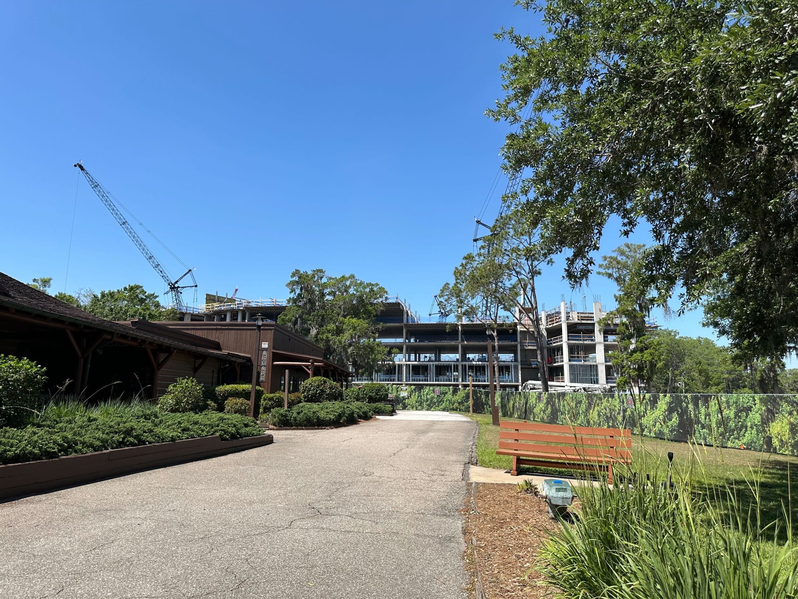 A construction site with a partially built structure and cranes in the background hints at the future Lakeshore Lodge. In the foreground, a paved pathway, green plants, and a wooden bench sit under a clear blue sky, setting the scene for what will soon be an inviting lakeside retreat.
