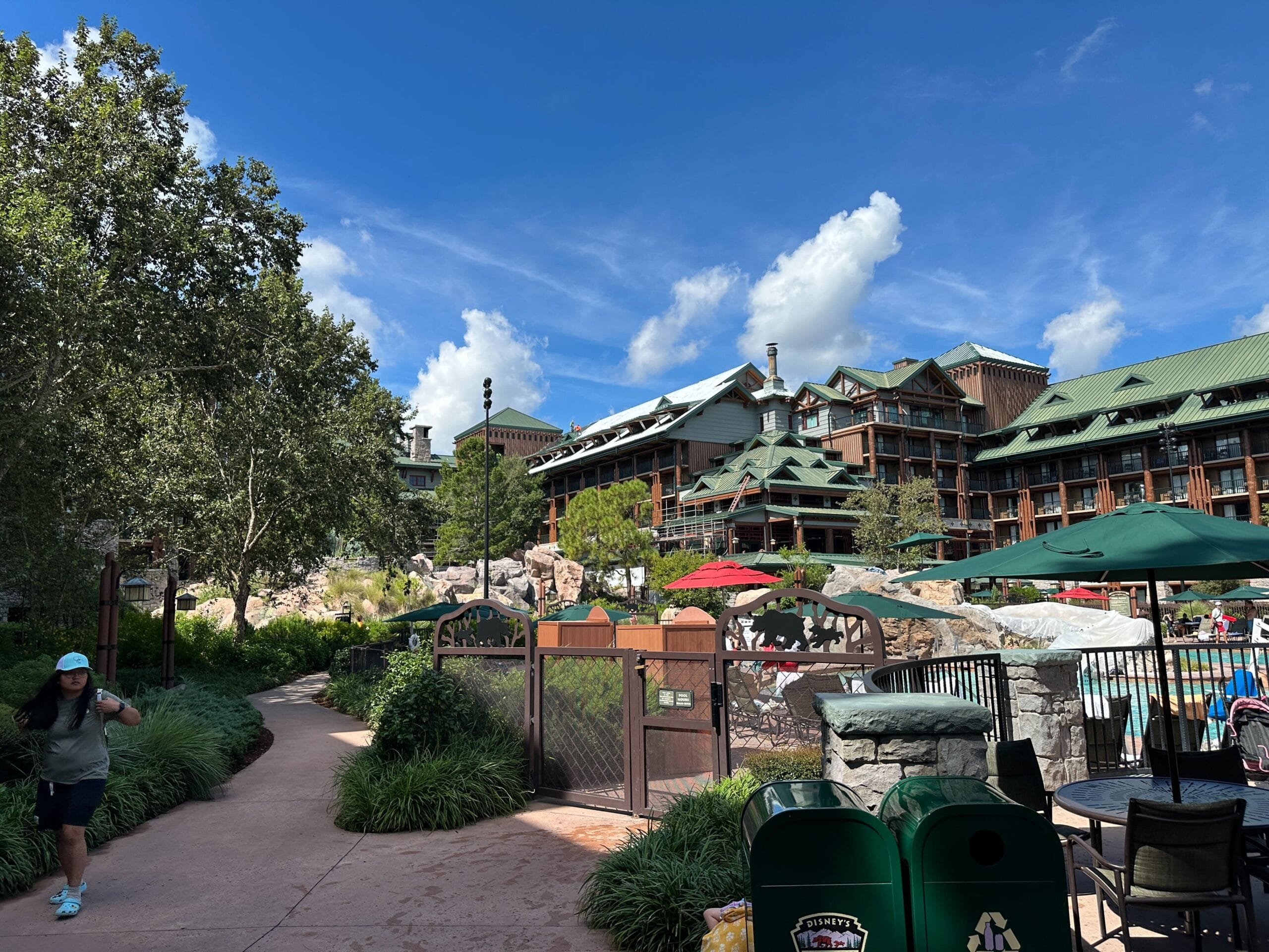 An updated Fort Wilderness Lodge resort hotel sits behind a gated outdoor pool with trees, umbrellas, and nearby walkers.