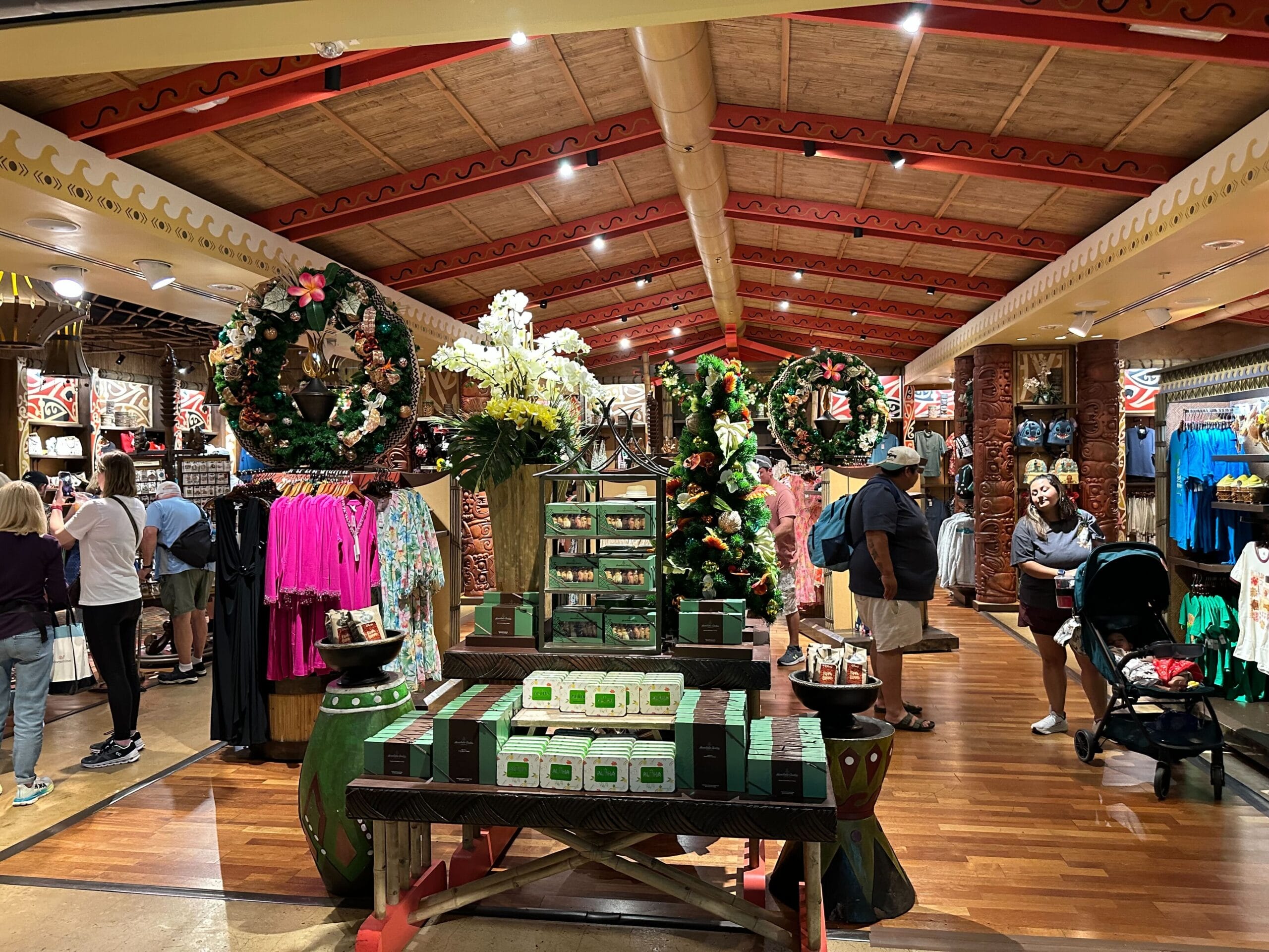 A bustling store with wooden beams displaying colorful clothing, Christmas wreaths, and various items on shelves and tables. Shoppers browse the merchandise.