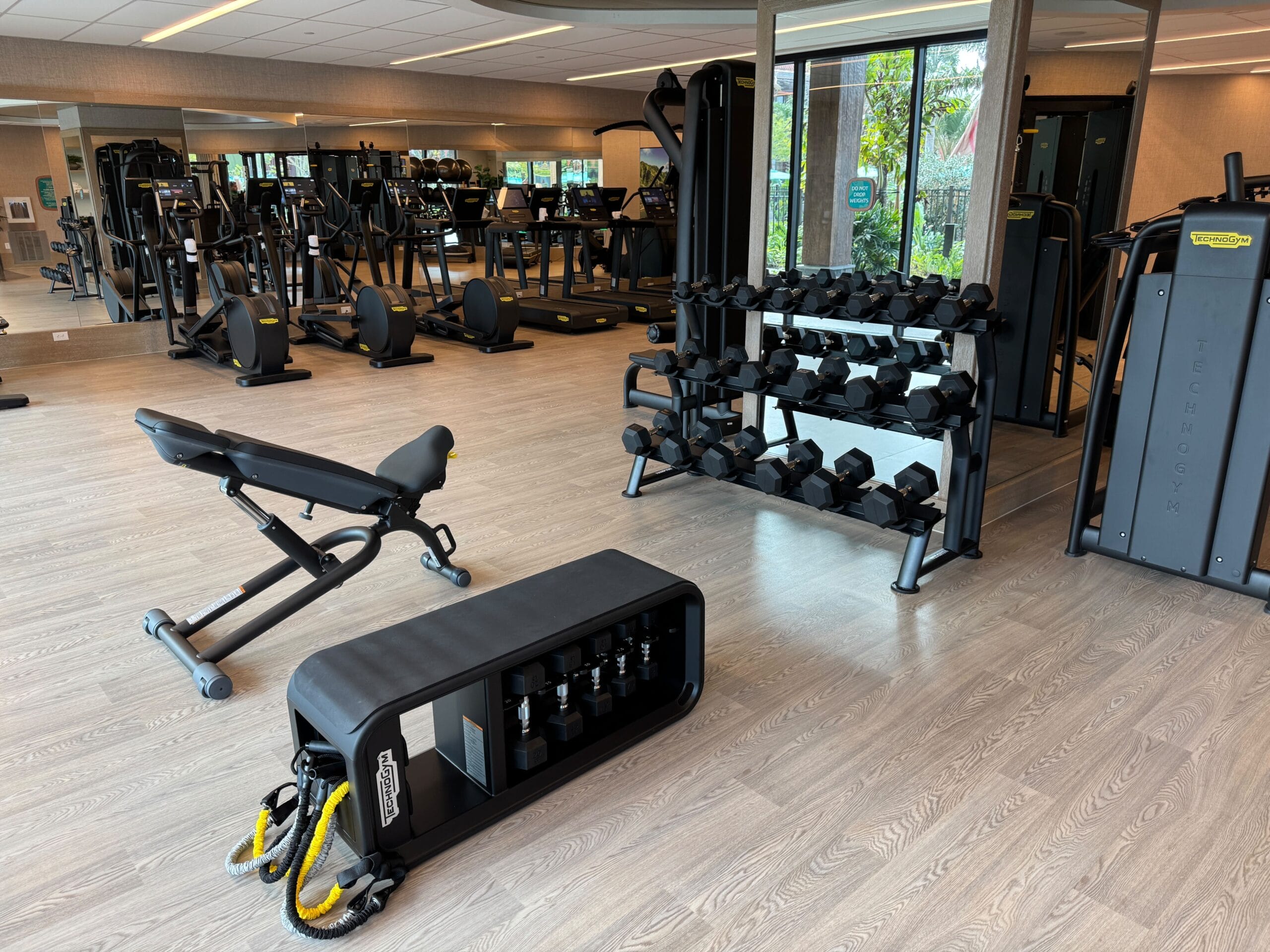 A modern gym with various exercise equipment, including weight racks, benches, cardio machines, and resistance bands, set on a wooden floor with mirrors on the walls.