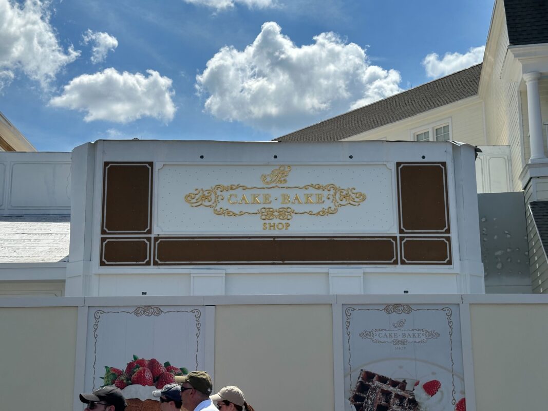 The photo shows the facade of The Cake Bake Shop with a decorative sign, against a partly cloudy sky. Several people stand in front of a barrier below the sign, seemingly captivated by the impeccable design that looks almost auto draft perfect.