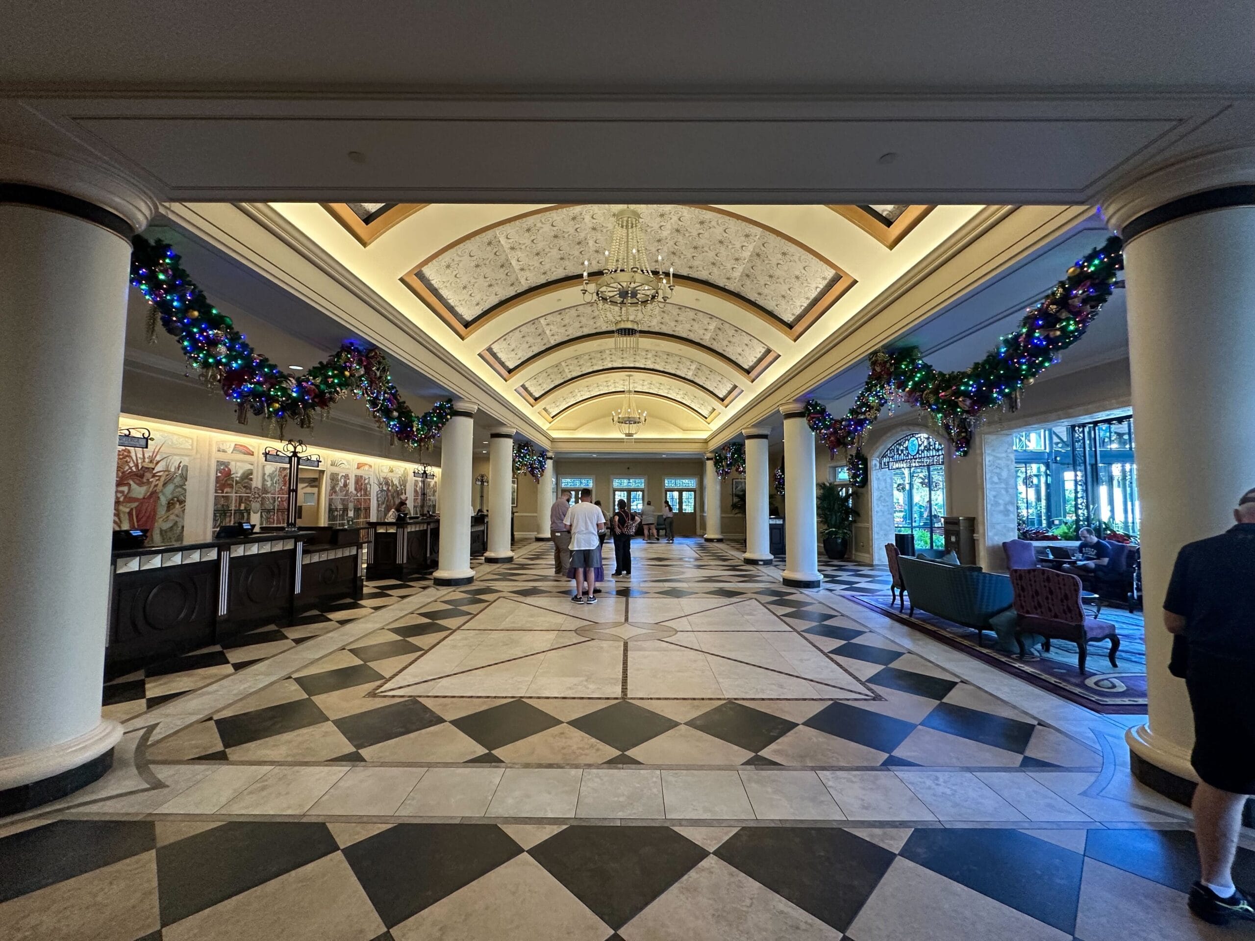A spacious hotel lobby decorated with garlands and festive lights. Elaborate ceiling, checkered floor, and seating areas create a welcoming atmosphere. Several people are present.