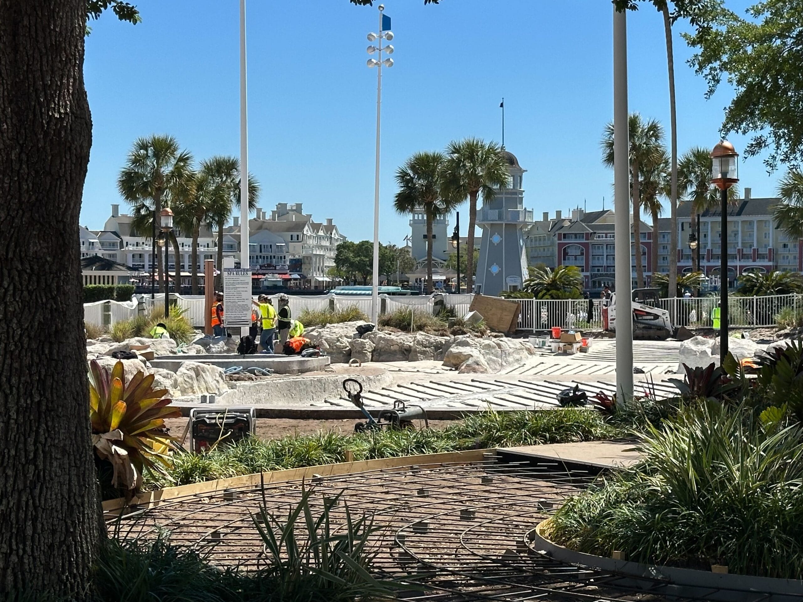 Amidst the construction site, workers diligently labor surrounded by palm trees and buildings, reminiscent of the bustling atmosphere near Disney's Yacht Club. Equipment and materials are scattered around under a clear blue sky, evoking a scene similar to the vibrant Stormalong Bay Pool.