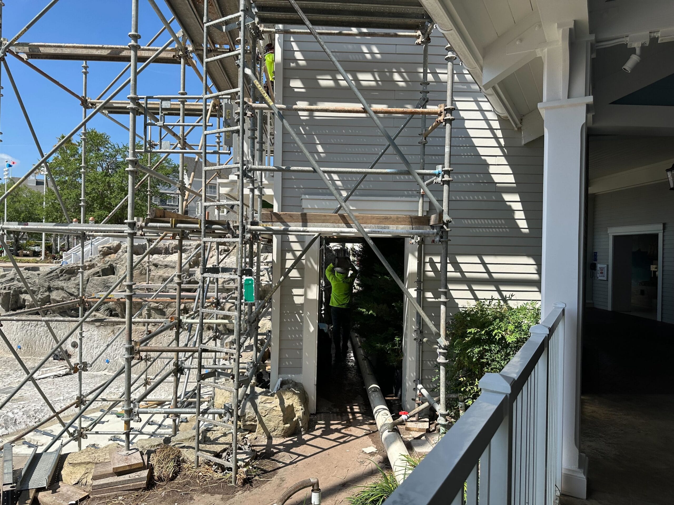 Scaffolding is set up around a building under construction, with pipes visible below. A worker in safety gear stands under the structure, reminiscent of the meticulous precision you'd find at Disney’s Yacht Club. A clear blue sky stretches endlessly above.