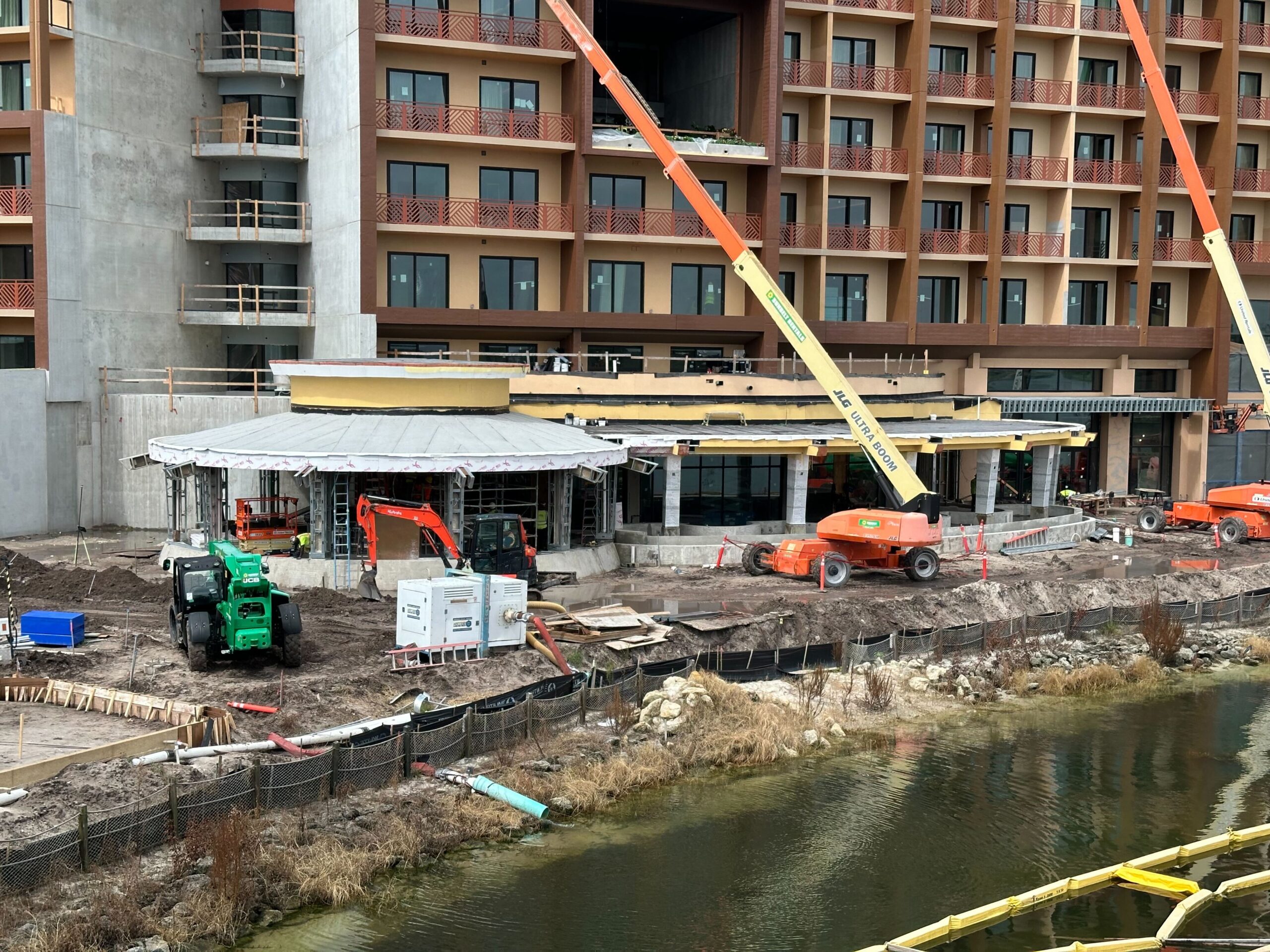A construction site near a building and a body of water, with machinery and workers. Several cranes and construction vehicles are present. The site appears to be in the early stages of development.