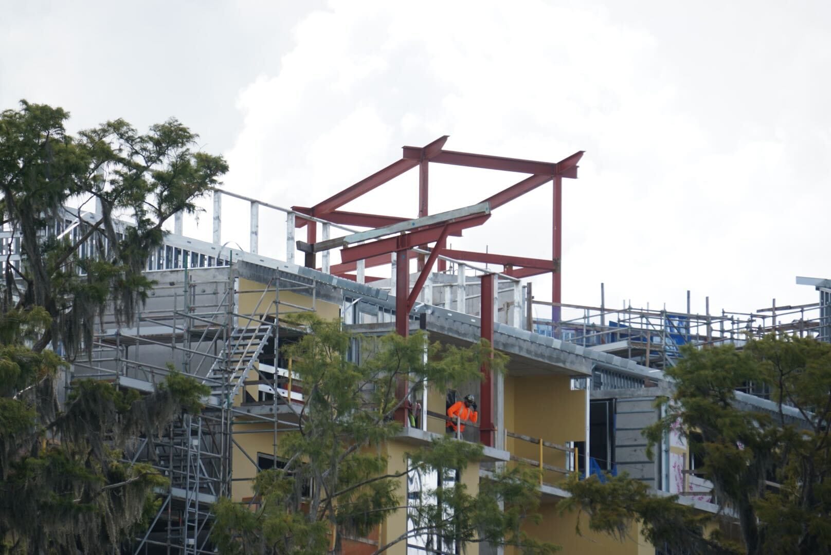 A construction worker in an orange vest stands on a partially built structure with exposed beams and scaffolding.