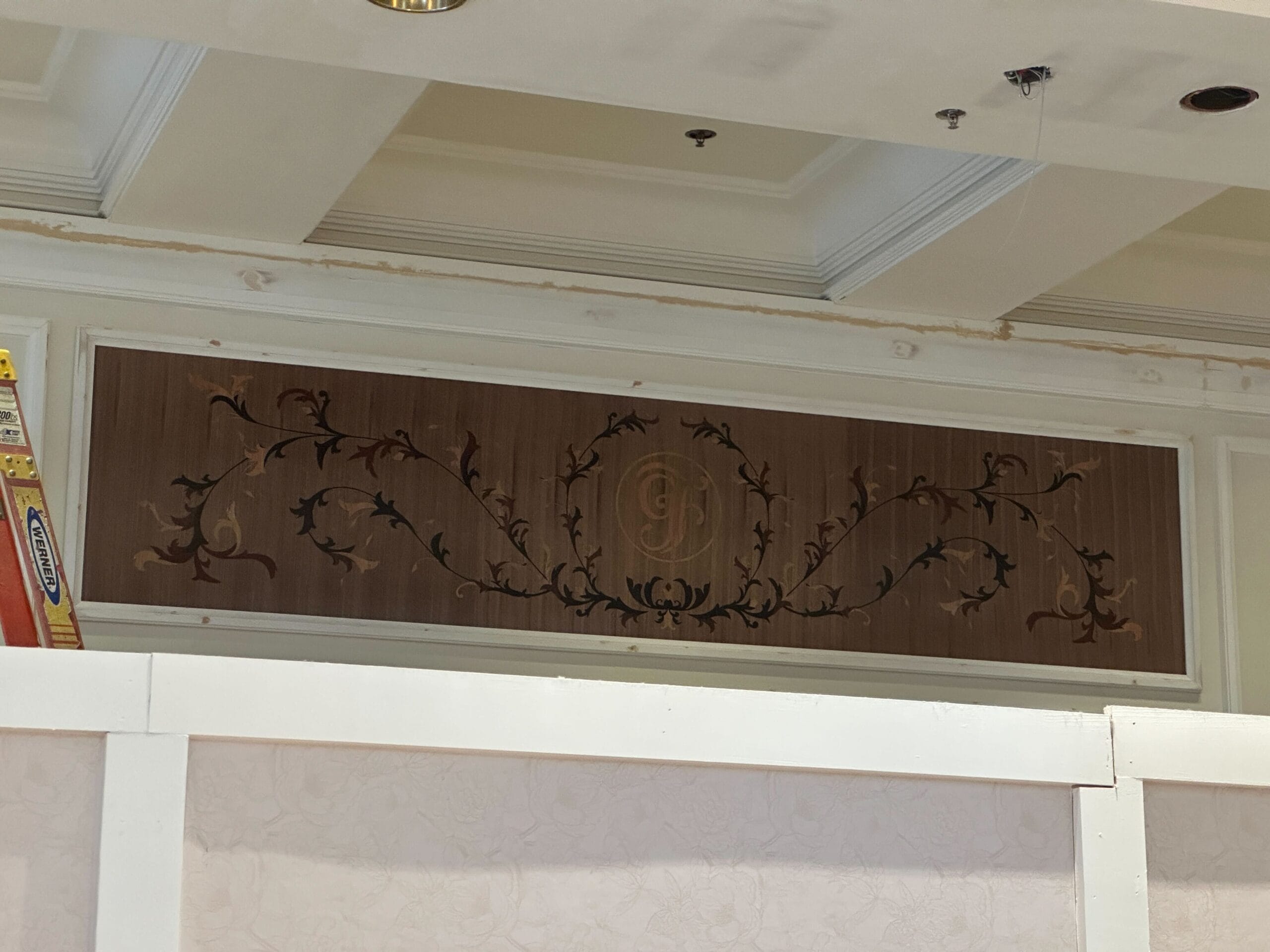 Decorative wooden panel with ornate scrolling vine designs and a central monogram above a white partition wall.