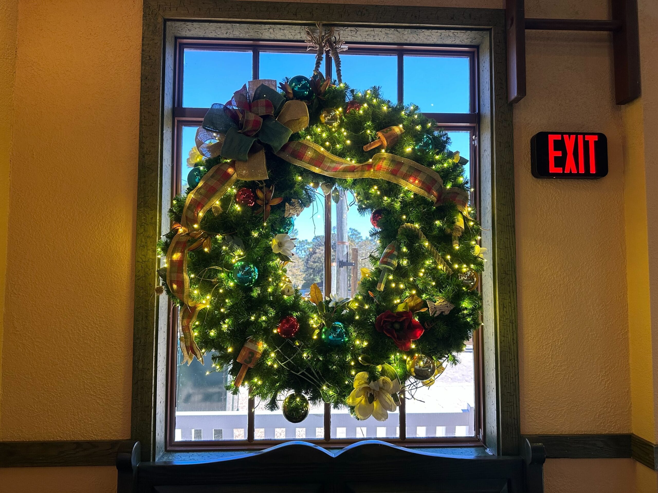 A large decorated wreath with lights hangs in a window next to an exit sign.
