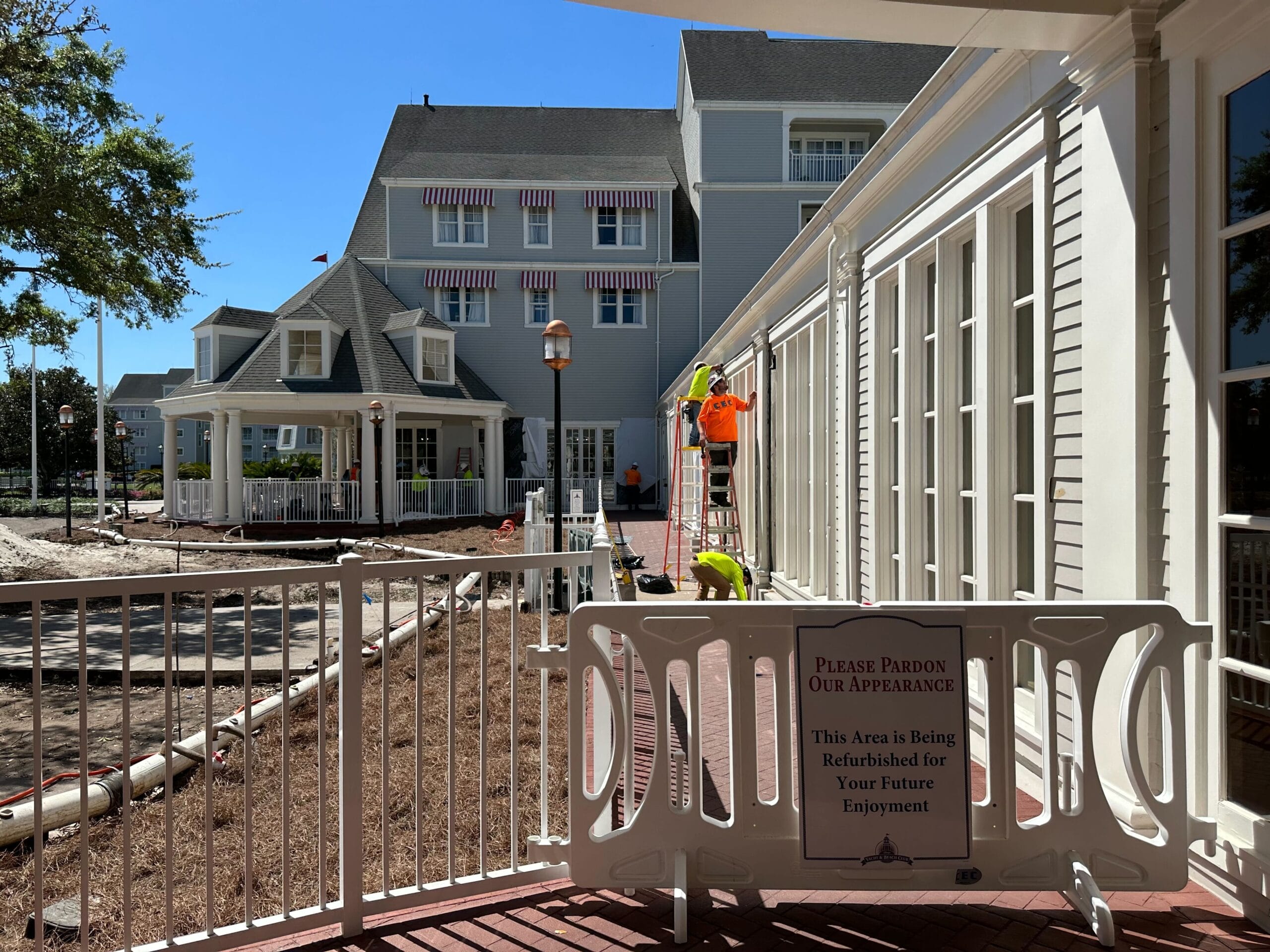 Exterior view of a large building under renovation, with workers wearing vests and ladders visible. A sign placed on a barricade reads, Please Pardon Our Appearance.
