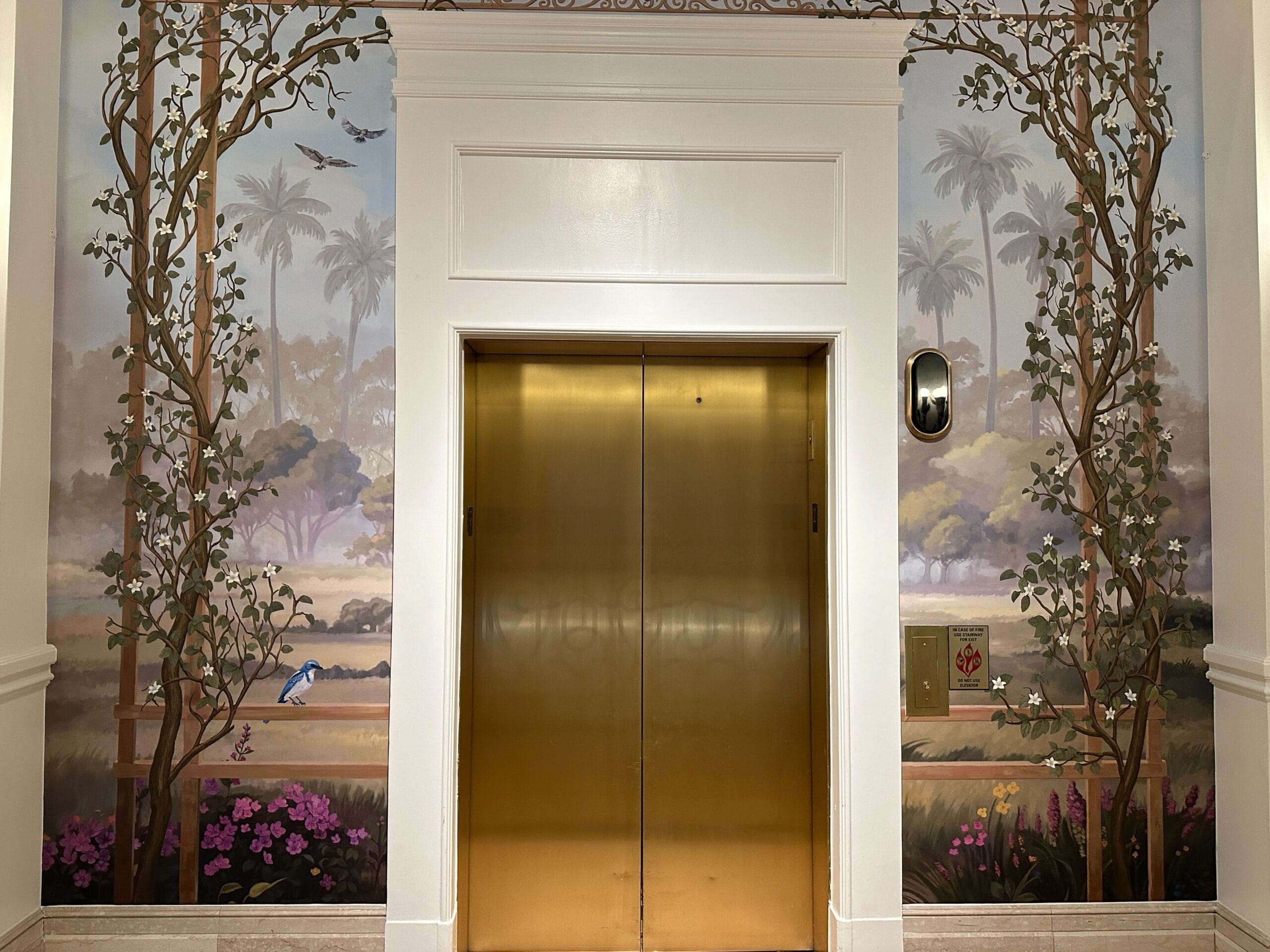 A gold elevator near bar countertops, decorated with a mural of tropical plants and birds, evokes the lush theming found at Disney resorts.