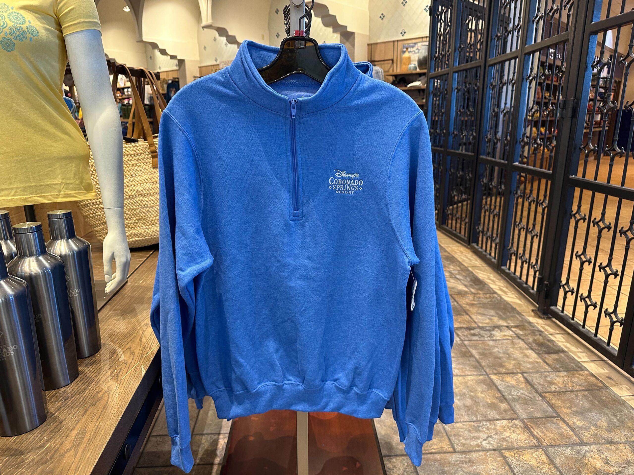 Blue quarter-zip sweater with Coronado Springs Resort logo displayed on a rack in a store, surrounded by metal water bottles and an array of other Coronado Springs merchandise.