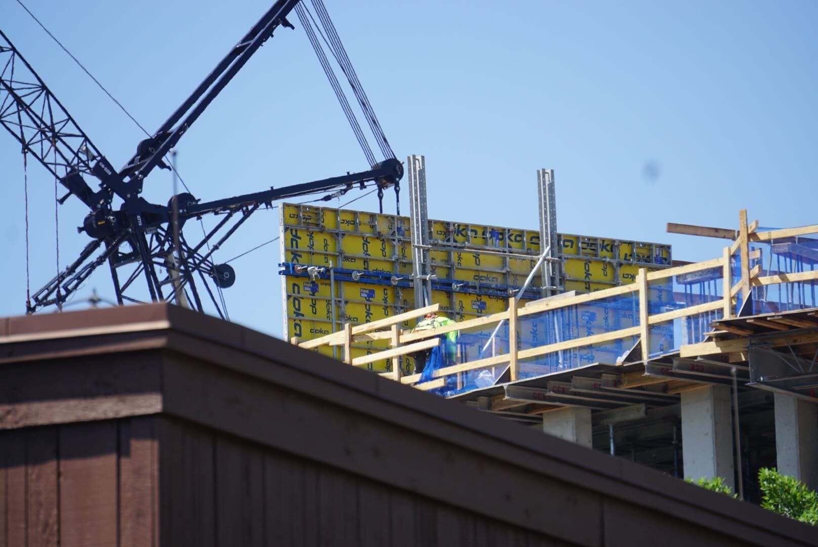 Under the clear blue sky, the Disney Lakeshore Lodge construction site buzzes with activity. A crane expertly lifts materials as yellow paneling and wooden scaffolding start to shape a towering dream. Stay tuned for your next exciting construction update!.