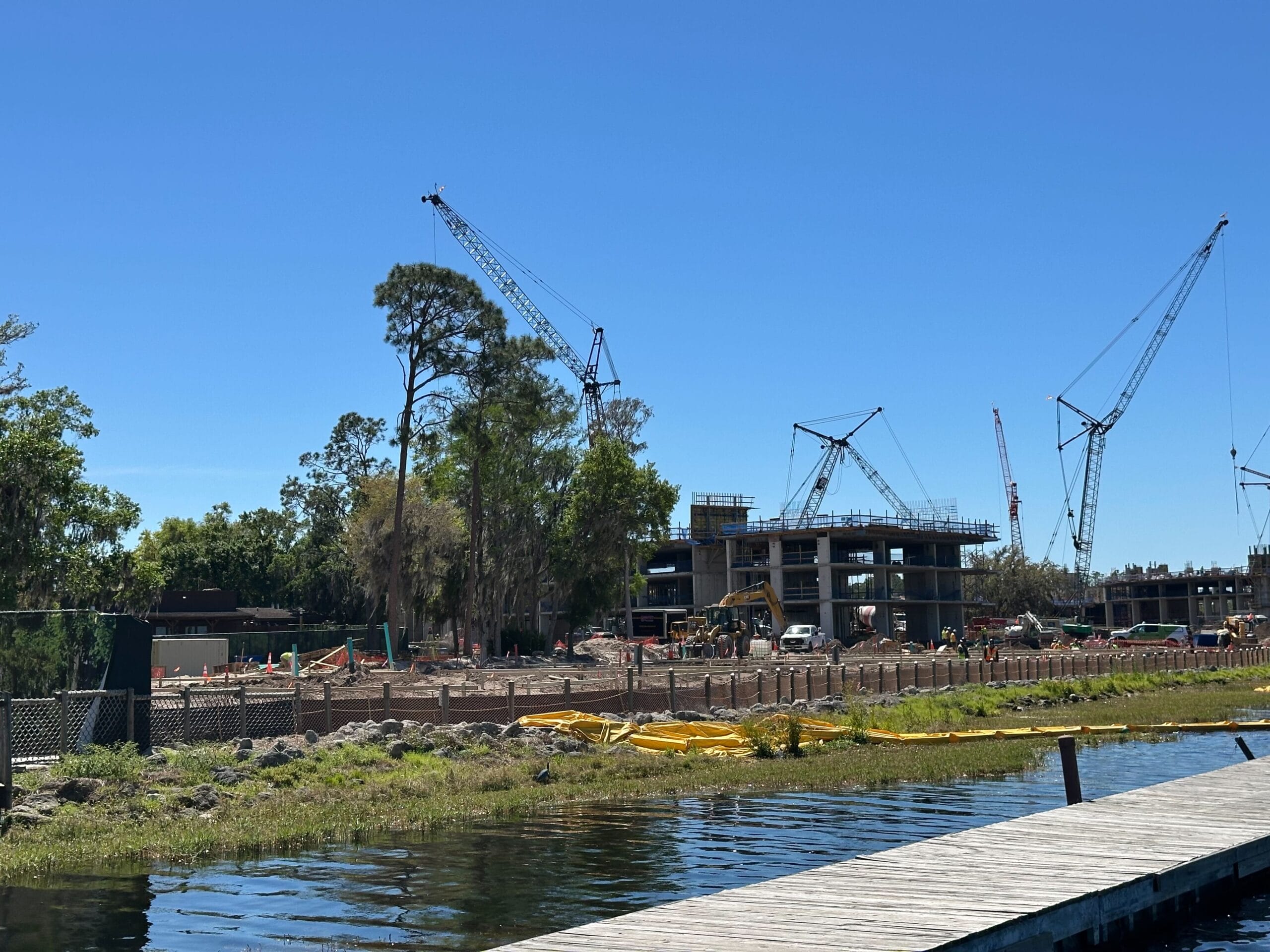 A construction site with cranes and partially built structures under a clear blue sky, surrounded by trees and bordered by a wooden dock and water.