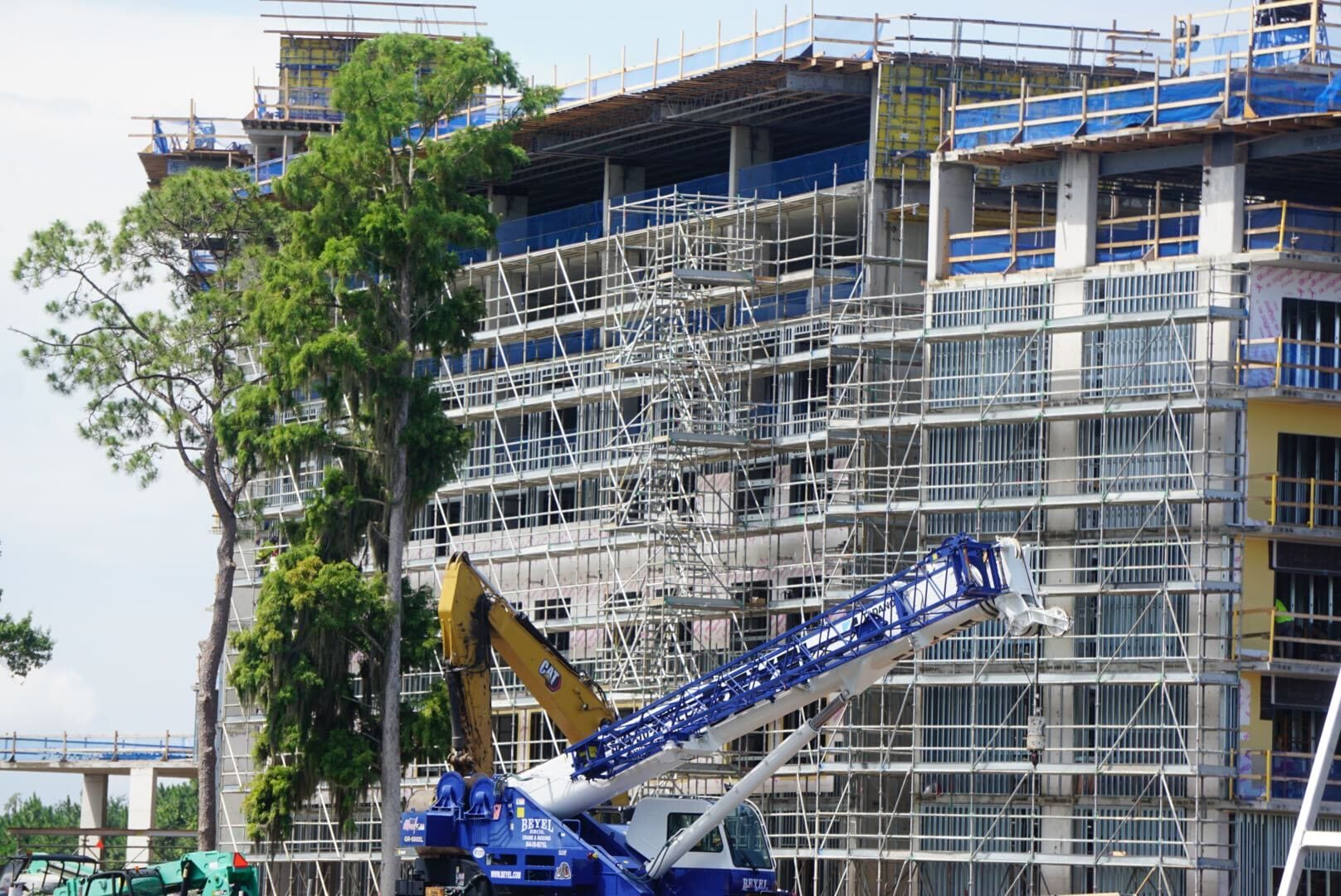 A large building under construction with scaffolding, a crane, and trees in the foreground.