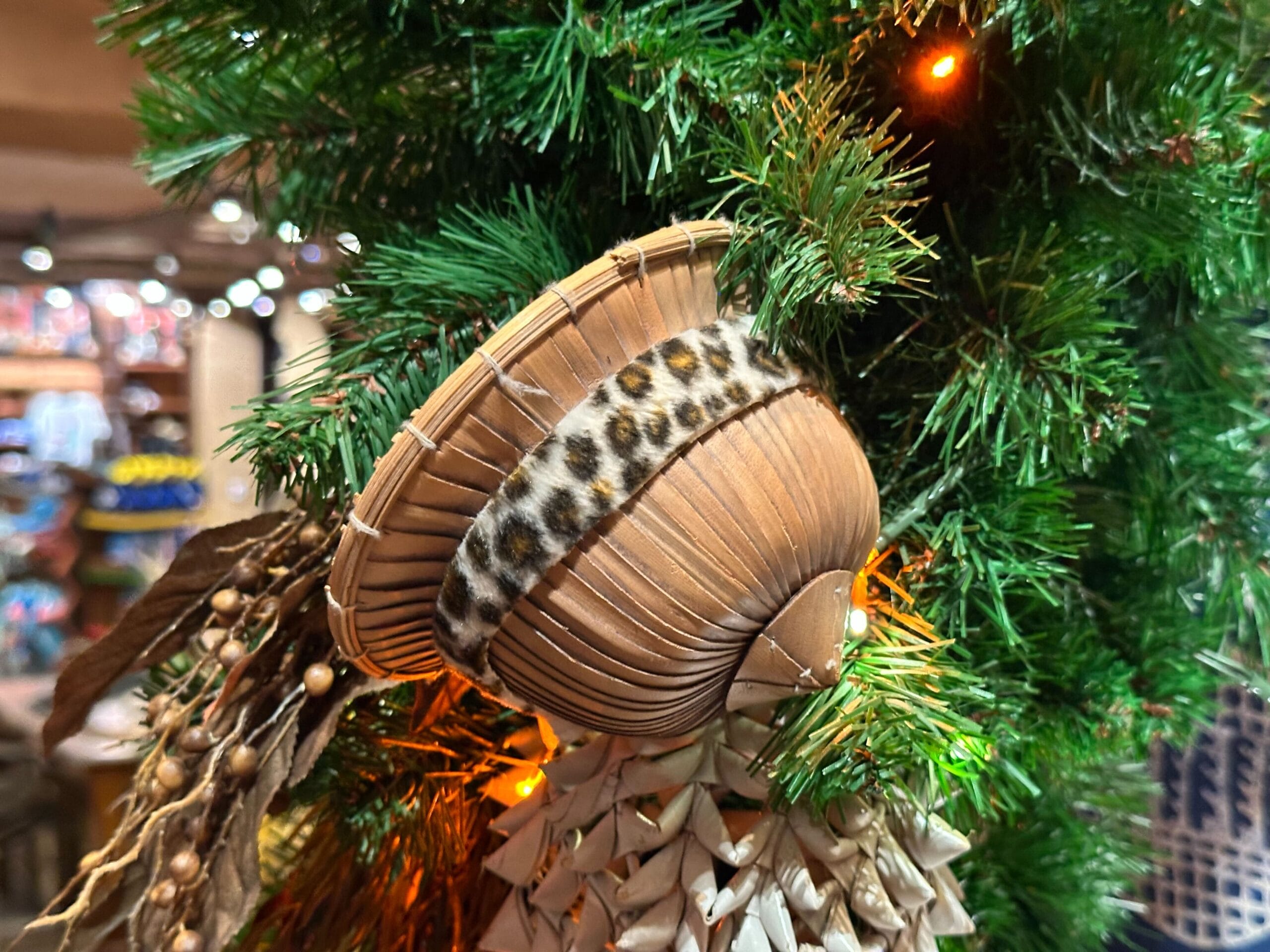 A decorative hat with a leopard print band hangs on a Christmas tree branch, surrounded by green foliage and soft orange lights.