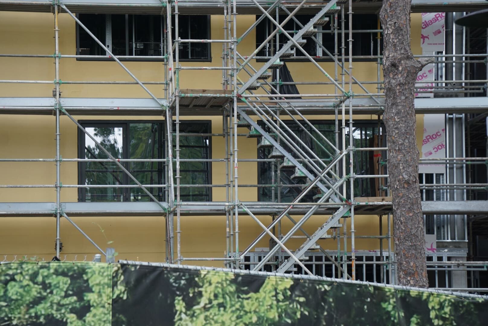 Scaffolding covers a yellow Lodge Construction building, with trees and a green printed barrier in the foreground.
