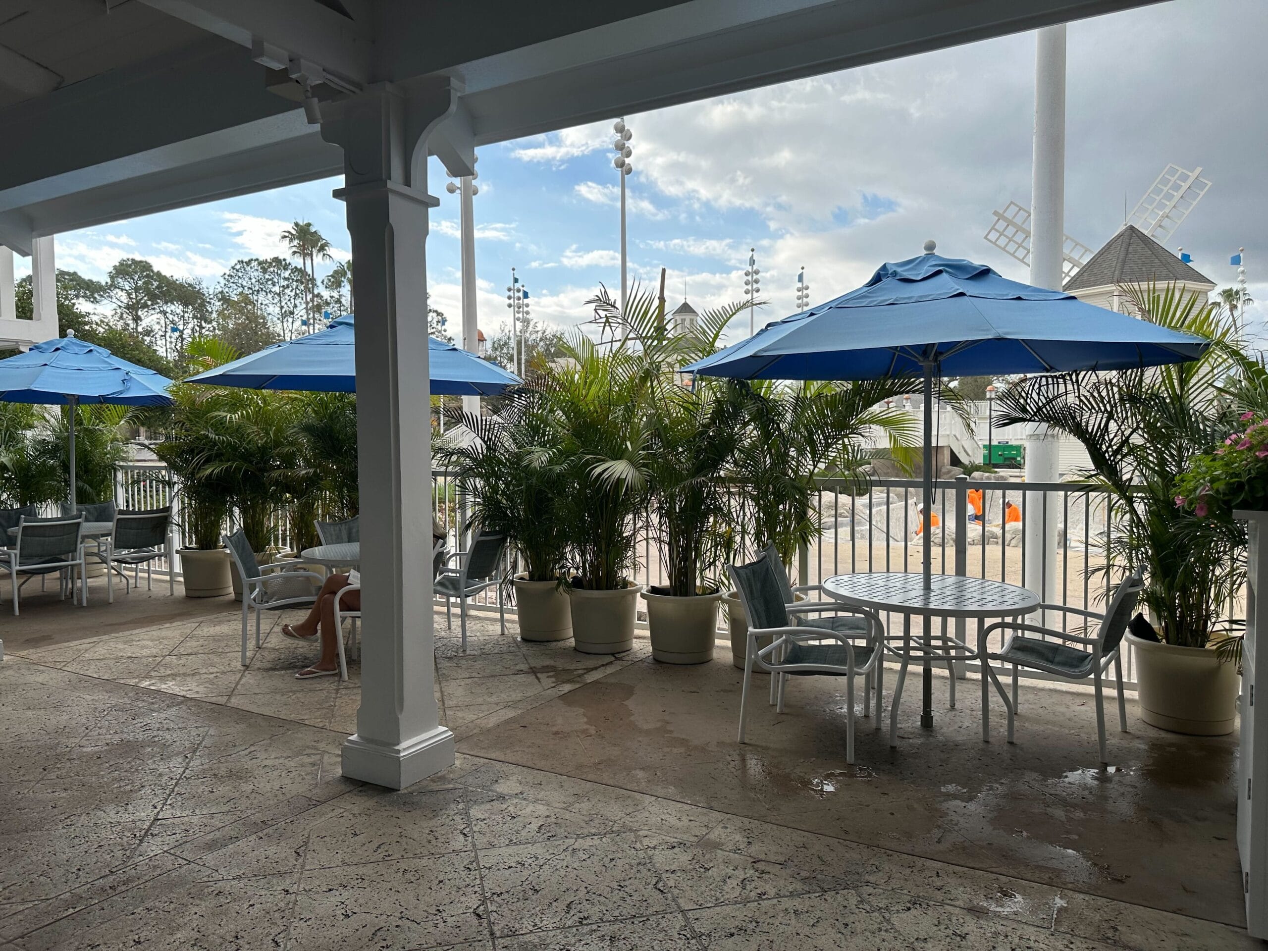 Outdoor patio with several tables and chairs under blue umbrellas. Potted plants surround the area, and a cloudy sky is visible in the background.