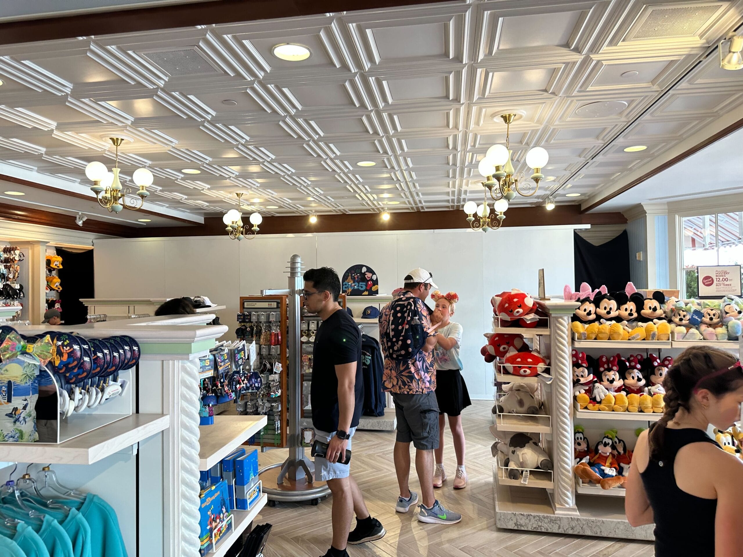 In the renovated section of Screen Door General Store at Disney's Boardwalk Inn, people browse plush toys and merchandise under a canopy of white ceiling tiles and chandeliers.