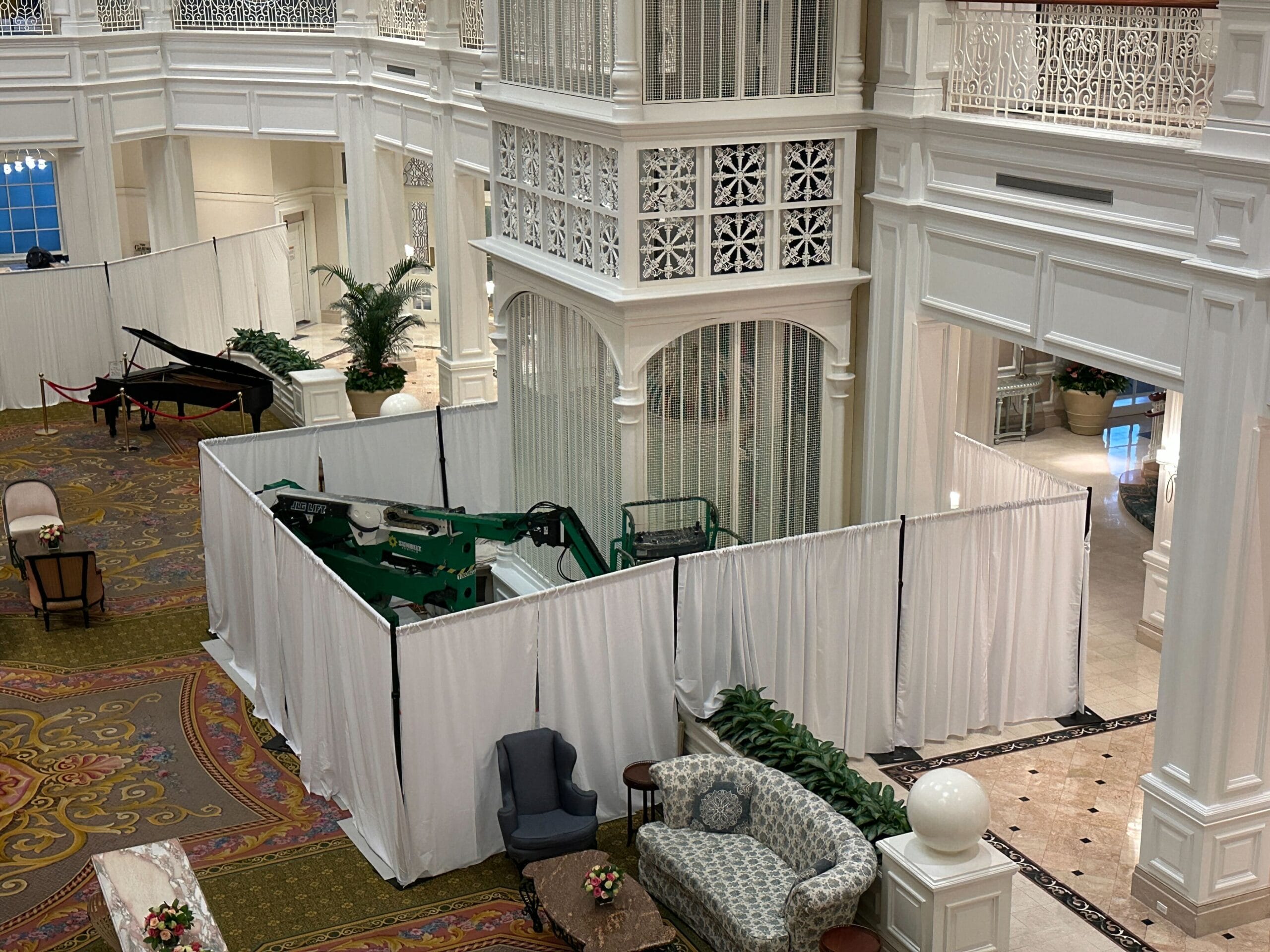 A white lobby with chairs and sofas, a piano, and a green lift behind white construction curtains.