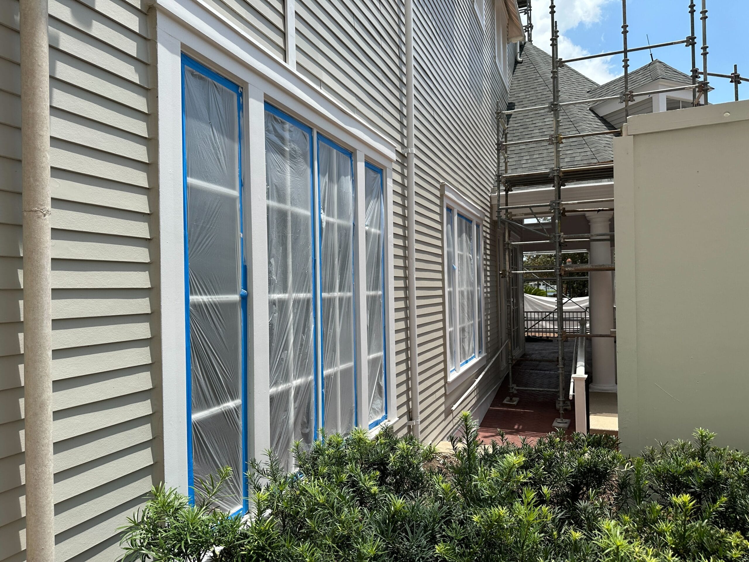 Large windows covered in plastic and blue tape on a house under renovation, with scaffolding and bushes nearby.