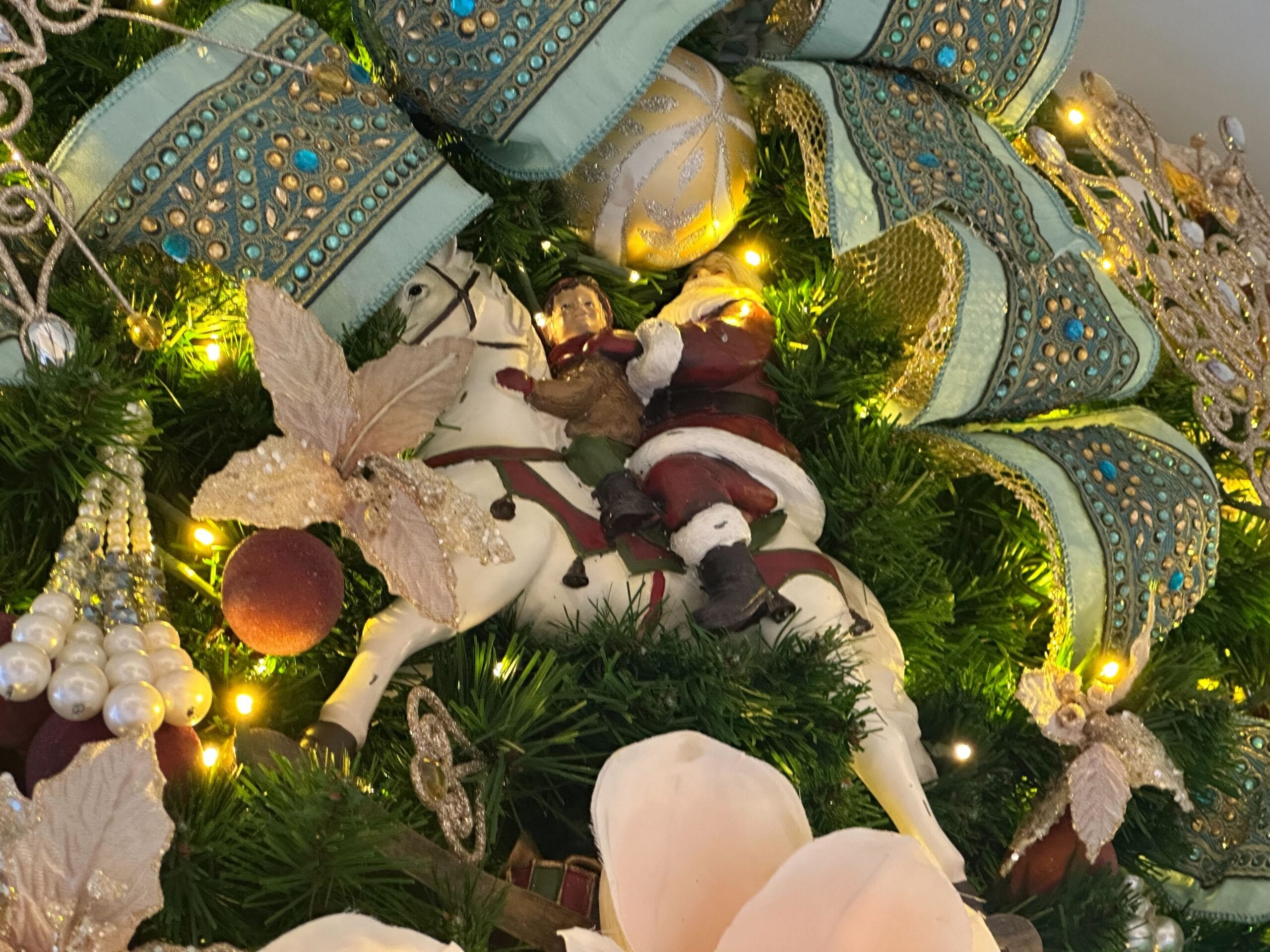 Close-up of a Christmas decoration featuring a Santa figurine riding a white horse, surrounded by ornate turquoise and gold ribbons and various festive ornaments with lights.
