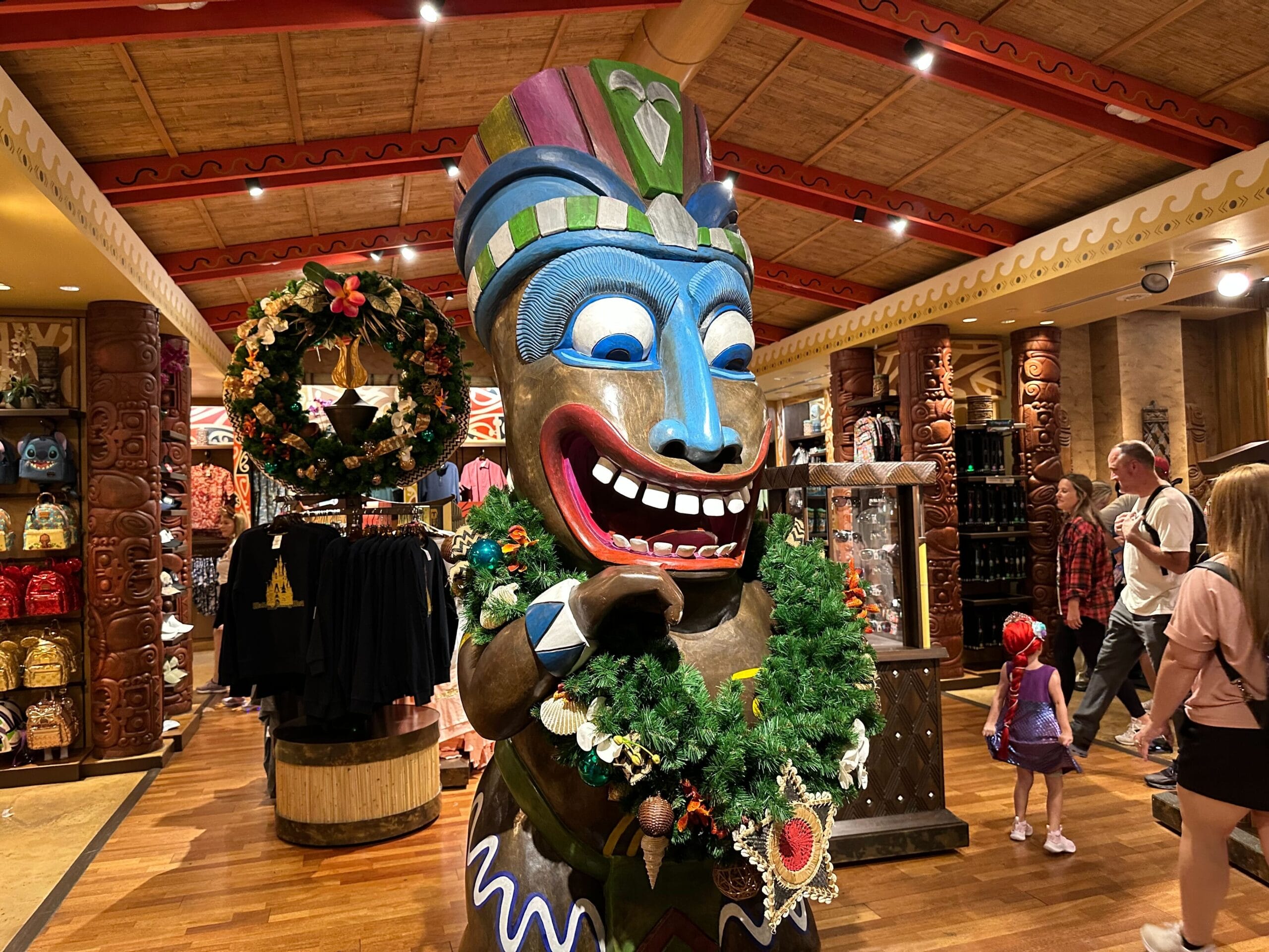 A colorful tiki statue adorned with a wreath stands in a store, surrounded by clothing and shoppers.