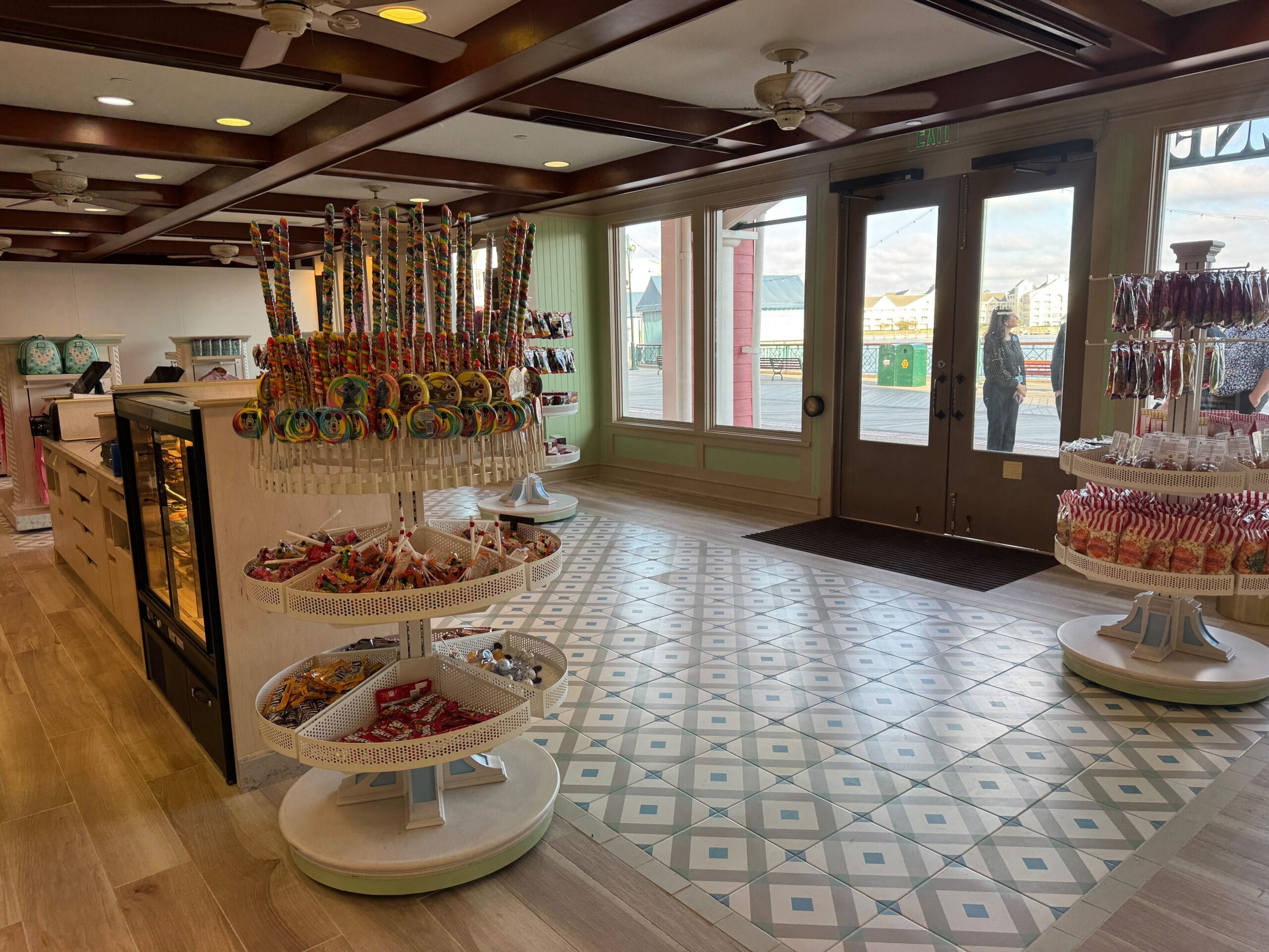 Candy shop interior with colorful lollipops on display stands, polished wooden counter, and patterned floor tiles. Large windows reveal an outside view.