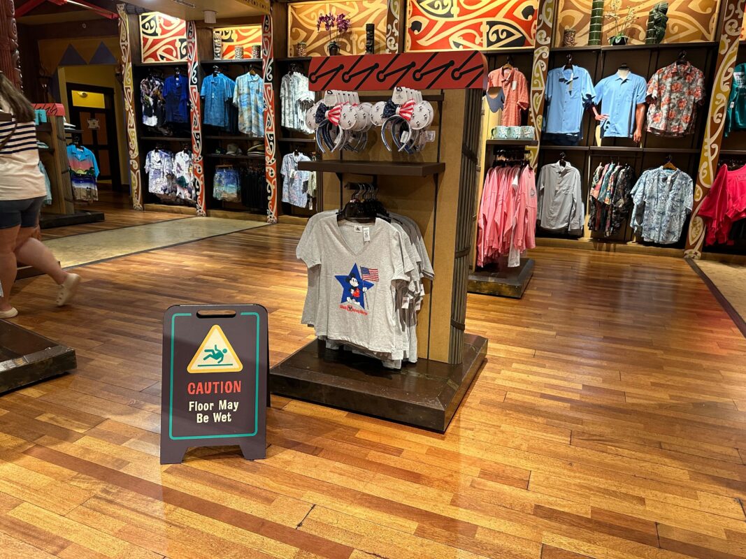 A clothing store with colorful shirts displayed, a wet floor sign in the foreground, and people shopping in the background.