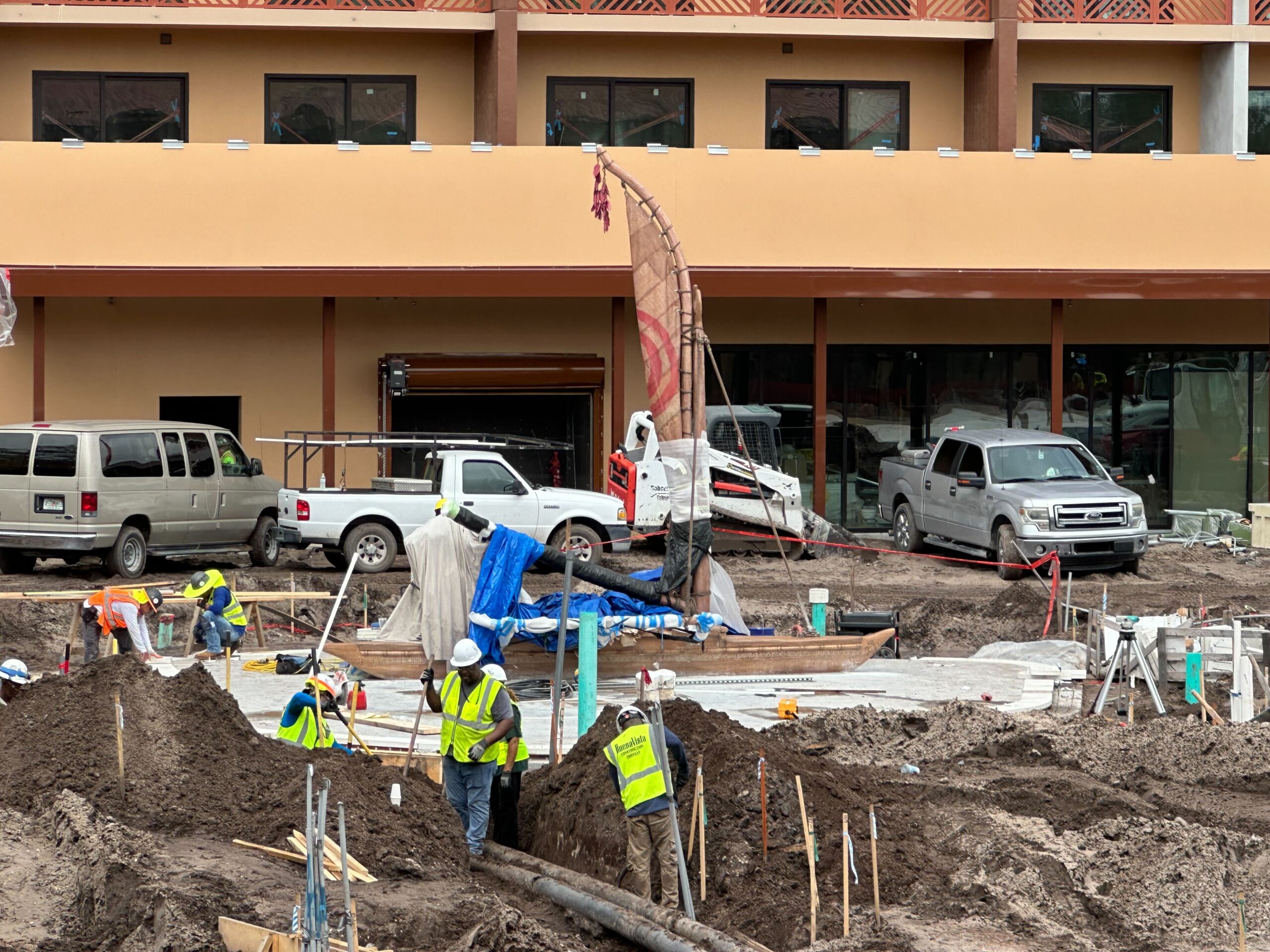 Several construction workers, wearing safety gear, work on a site with various equipment and vehicles in front of a building with large windows and balconies.
