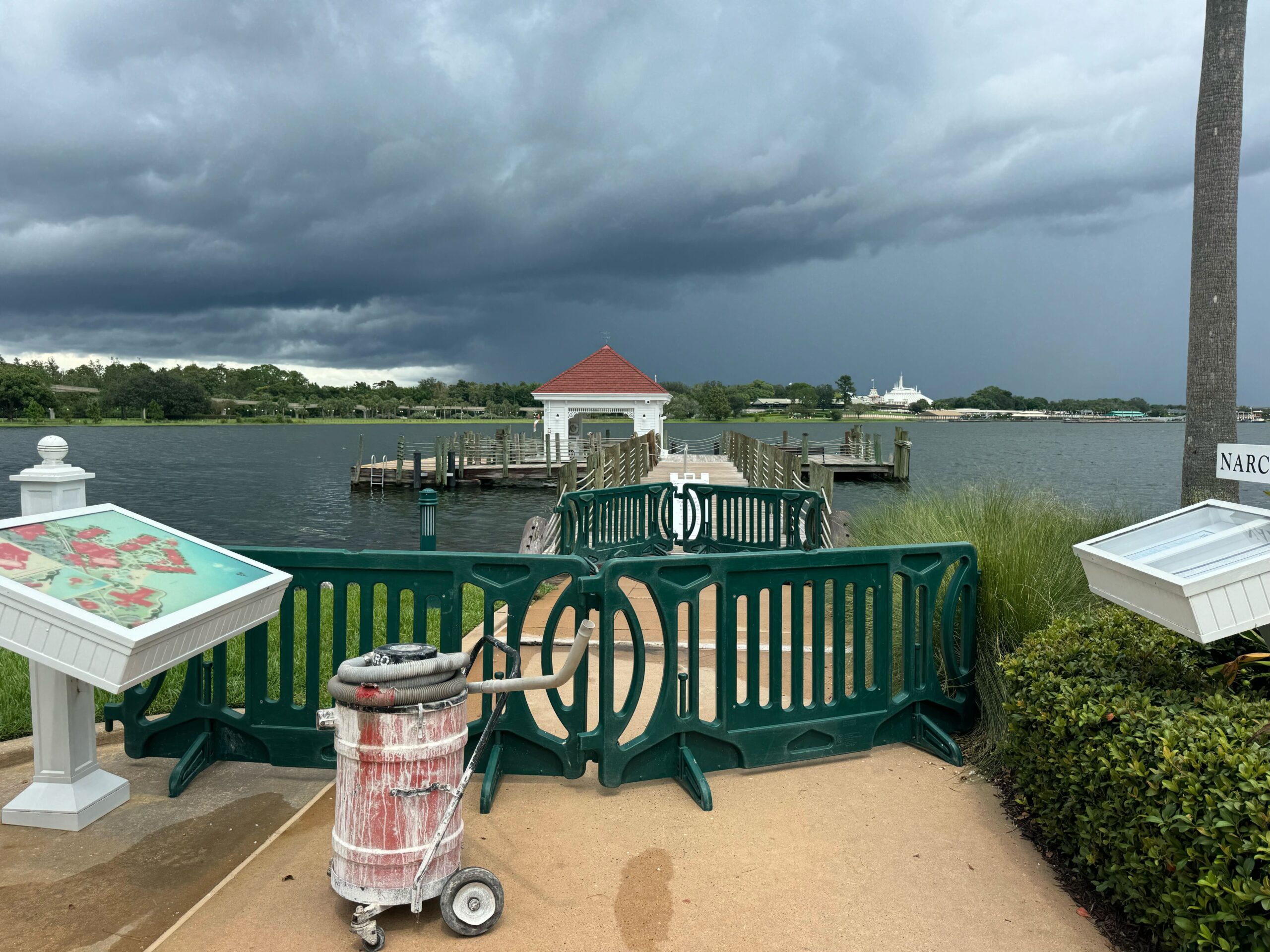 A small dock area by a lake, surrounded by green barriers and a cart with painting supplies in front. Dark storm clouds form over the horizon. Nearby signage displays maps and information.
