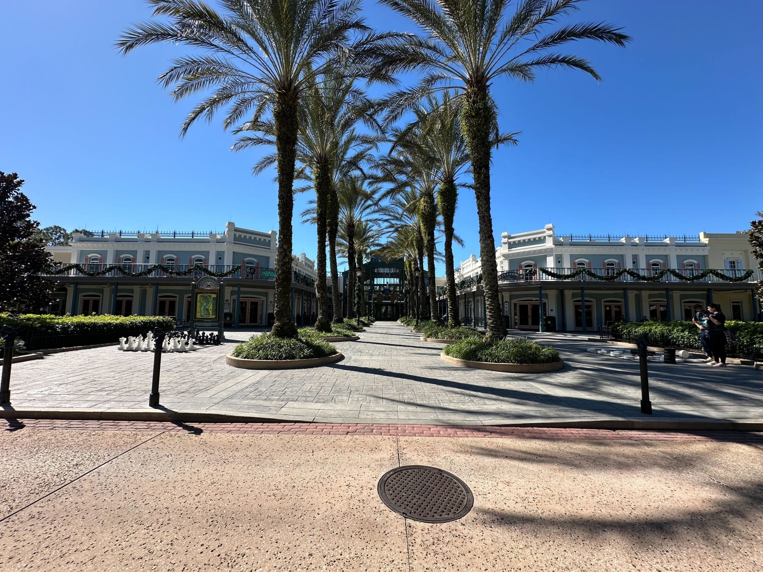 A wide pathway lined with palm trees leads to a large building under a clear blue sky. The building is flanked by smaller structures on either side.