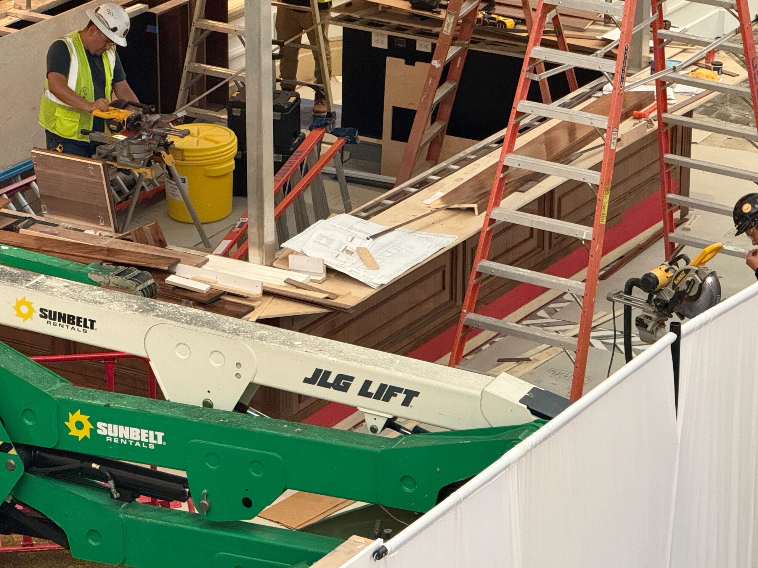 A construction worker uses a saw near ladders and lift equipment at an indoor building site with wood and plans visible.