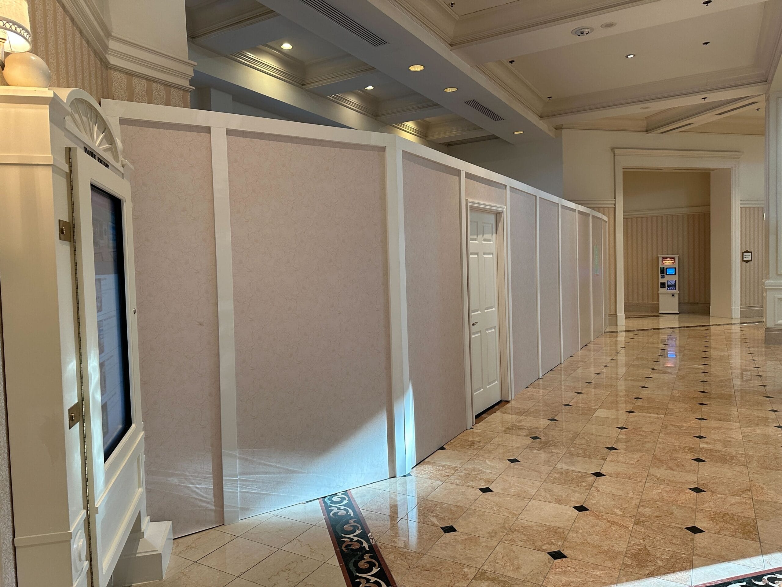 A white temporary wall with a closed door runs through a polished hallway with tiled floors and decorative ceilings.
