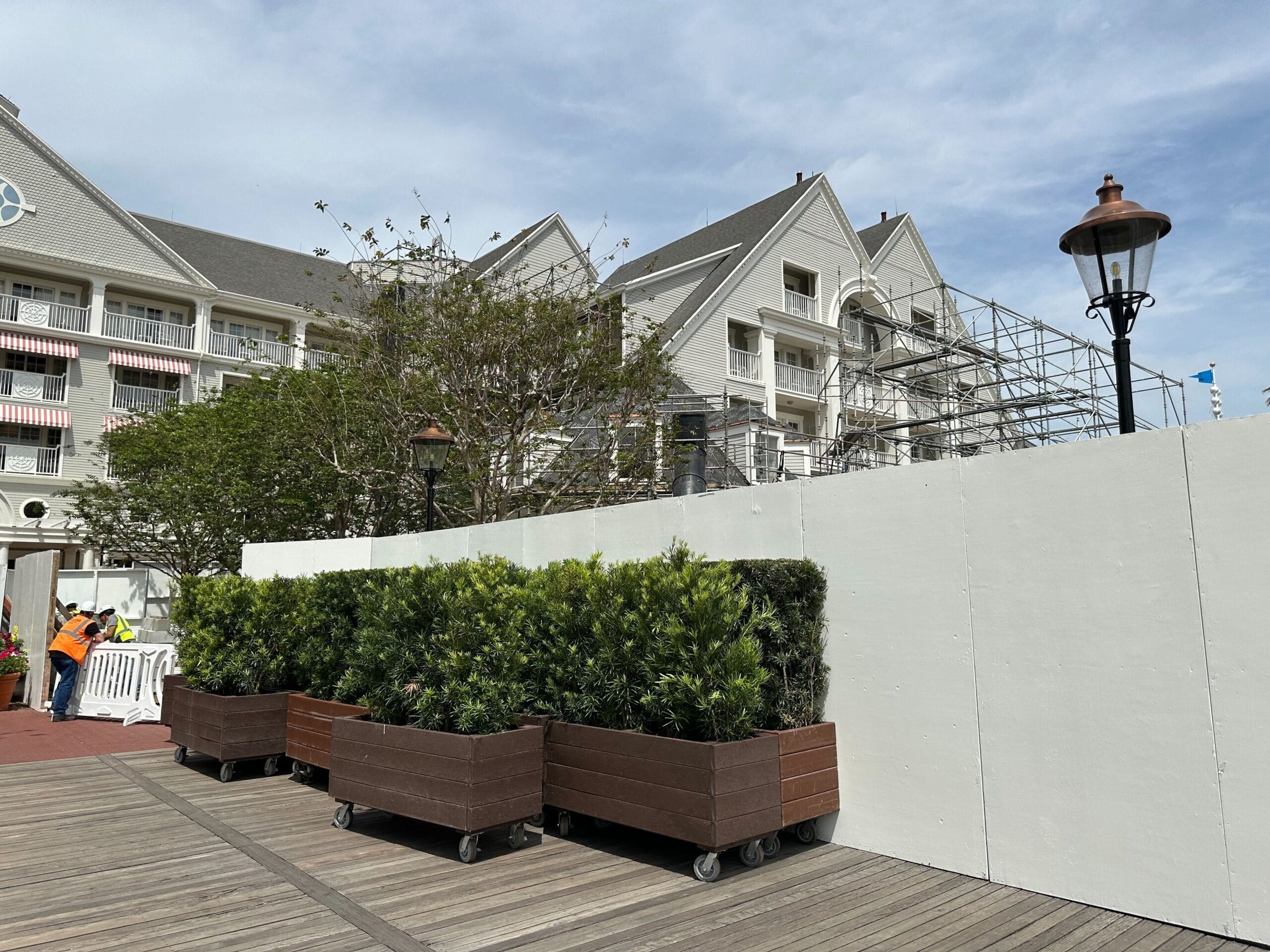 A building under renovation, its scaffolding visible alongside a white construction barrier. Amid planters brimming with greenery and two workers in safety gear on the wooden boardwalk, the scene evokes the charm of Disney's Yacht Club.