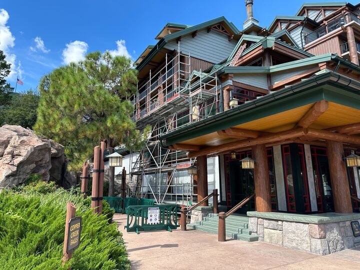 A Fort Wilderness Lodge-style building with scaffolding and construction signs, surrounded by greenery under a blue sky.