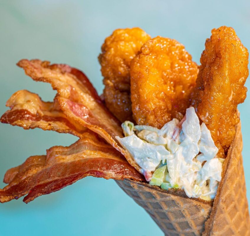 A waffle cone filled with fried chicken strips, strips of bacon, and coleslaw, perfect for enjoying at Typhoon Lagoon's Disney H2O Glow After Hours against a light blue background.