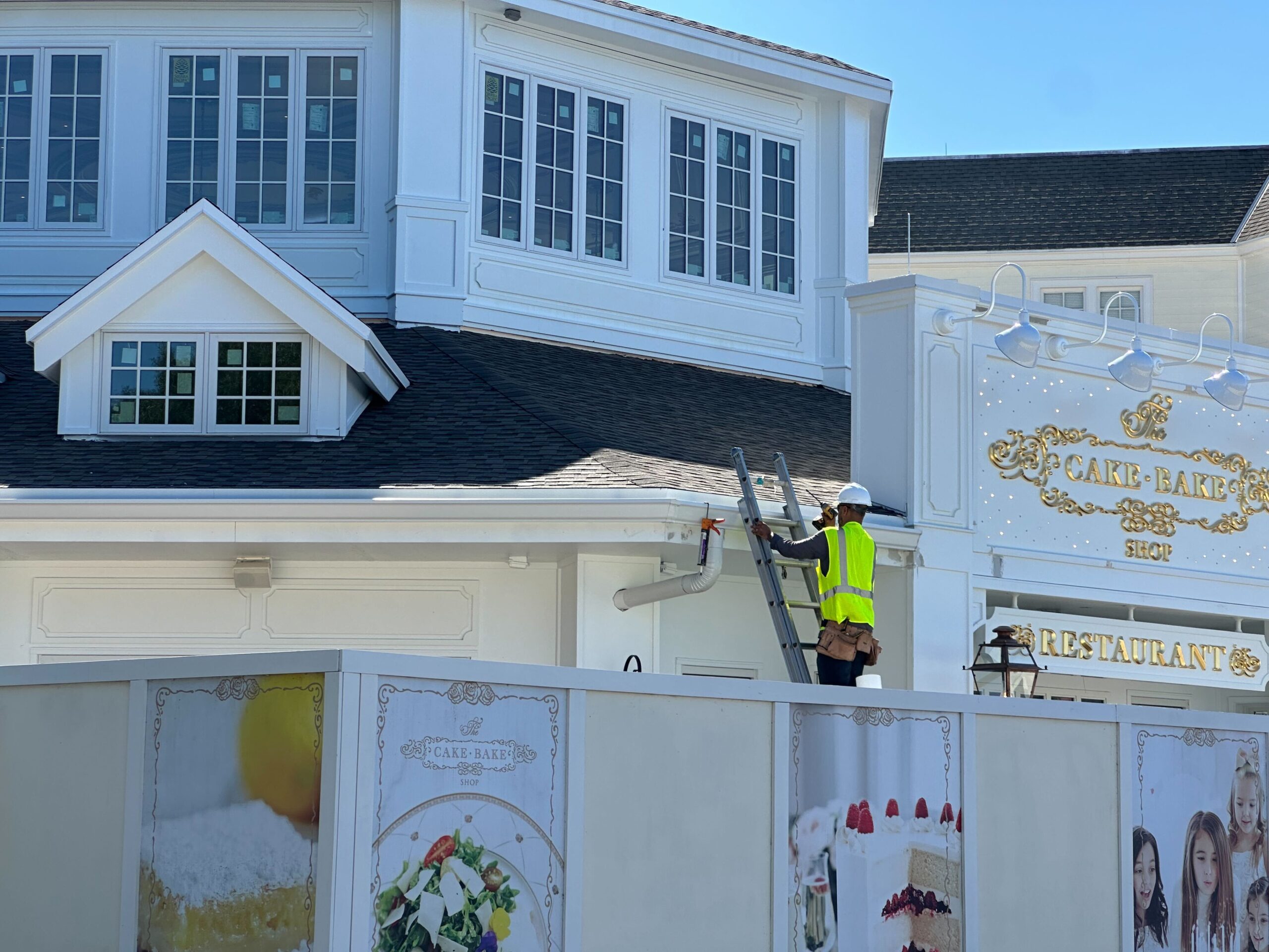 A construction worker in a yellow vest and hard hat climbs a ladder to a white building, with a decorative restaurant sign in the background. There is a white construction barrier in the foreground.
