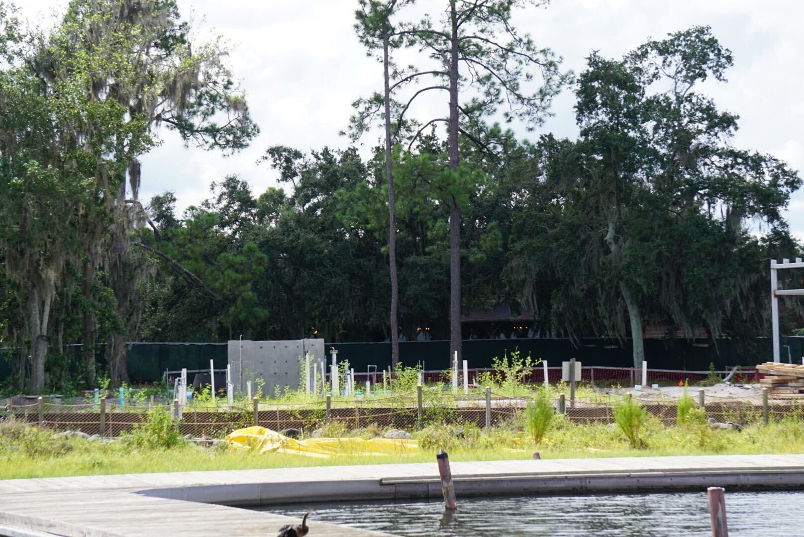 A fenced-off construction site with scattered pipes and materials, trees in the background, and a dock in the foreground.